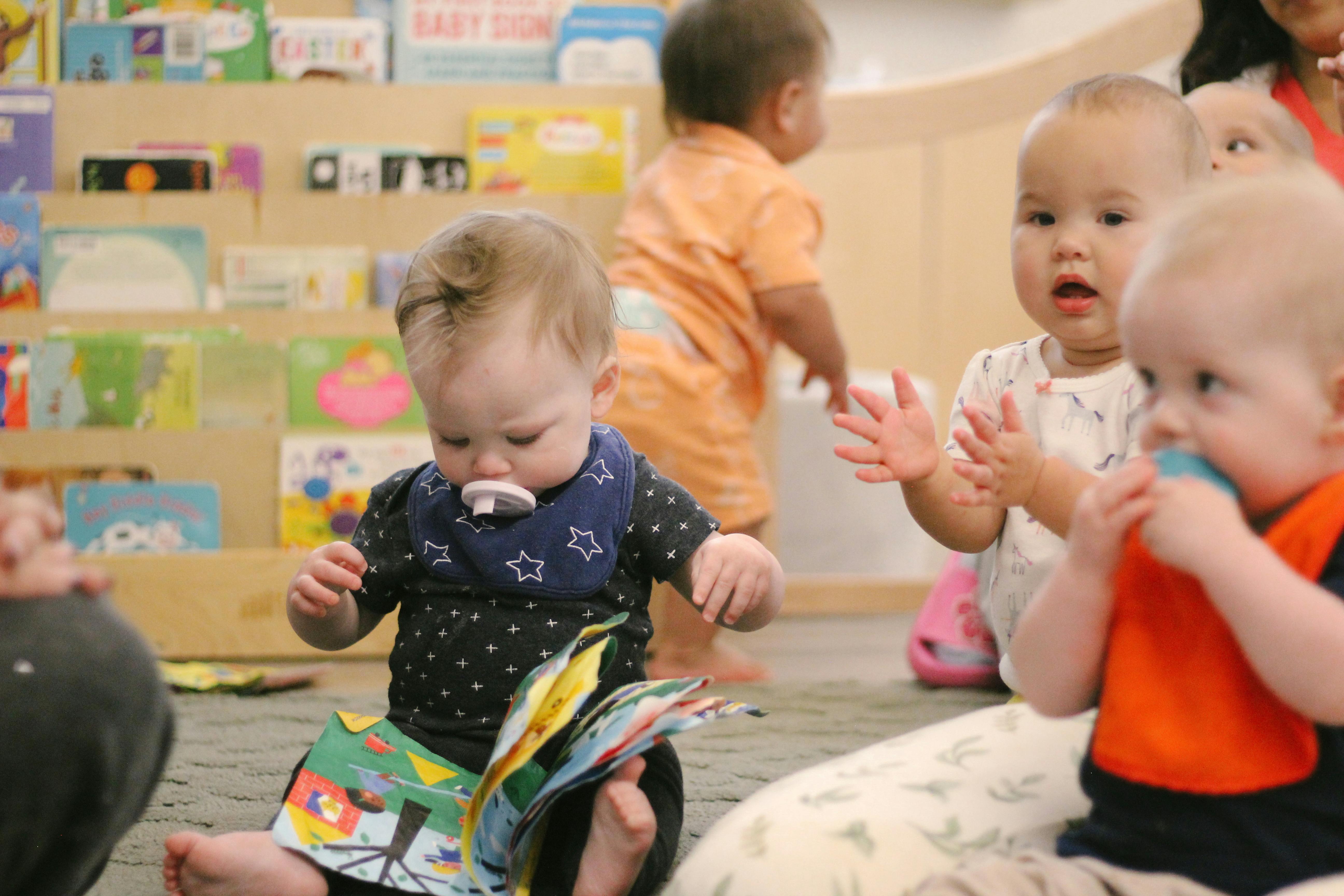 Child with a pacifier looking at a book, other children in a classroom