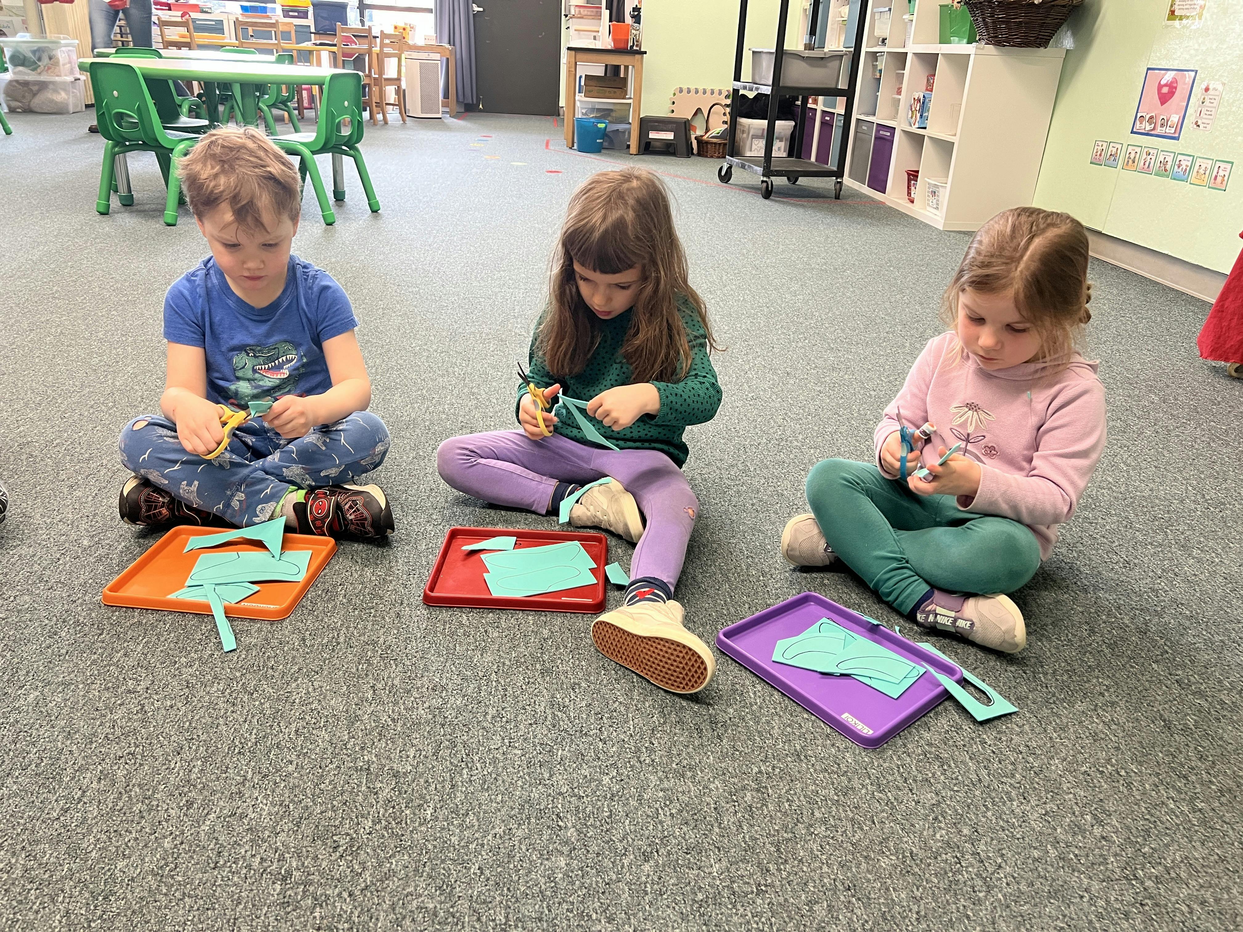 Three children sitting on the floor cutting paper shapes with scissors