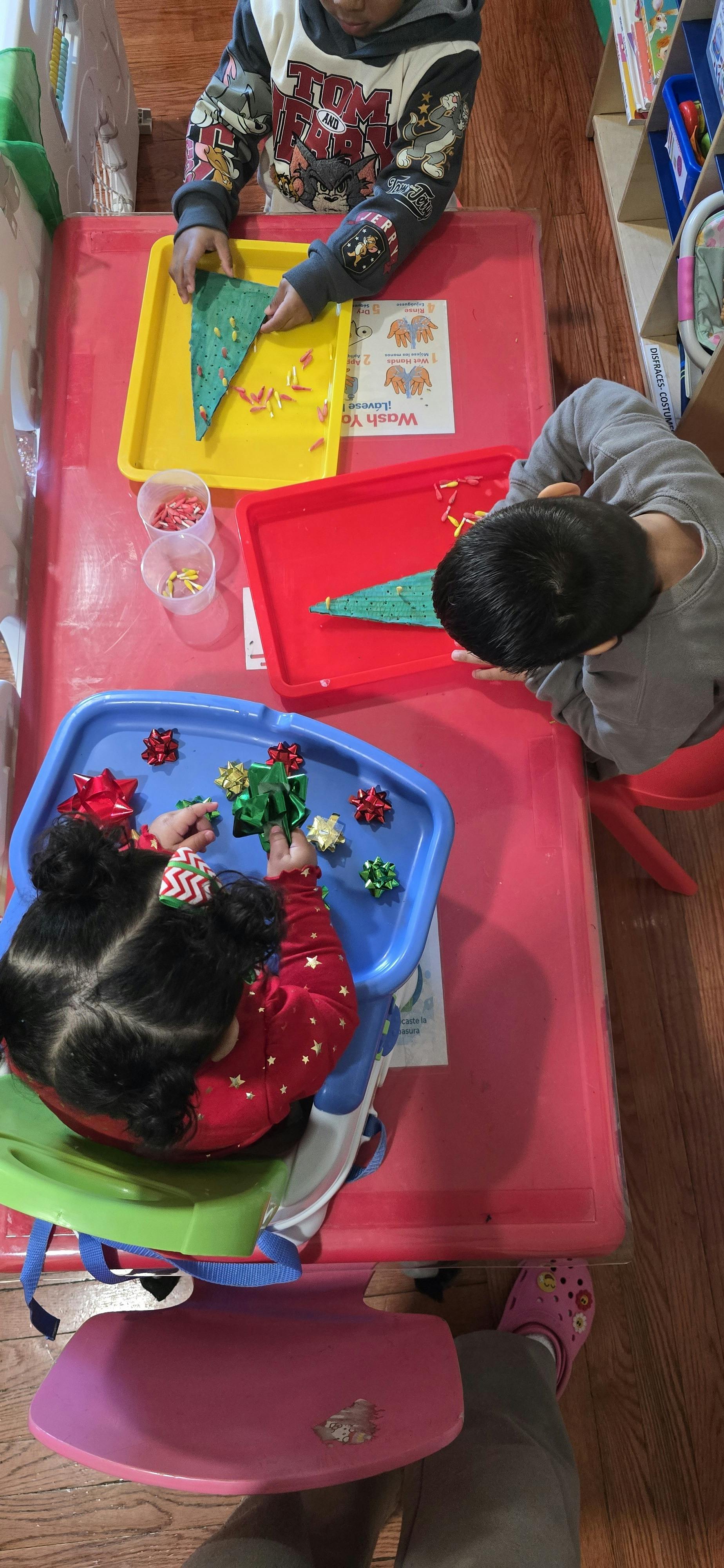 Three children crafting at a red table, decorating green triangles and sorting bows