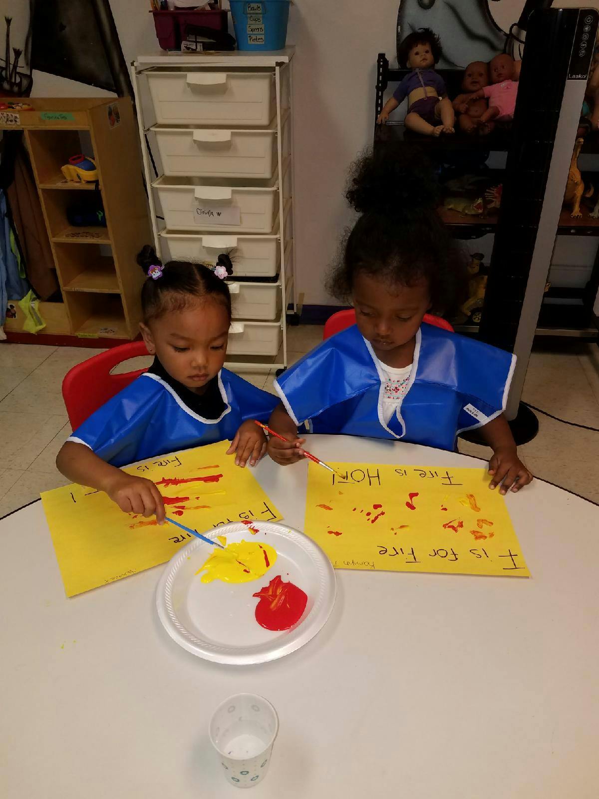 Two children in blue smocks painting on yellow paper at a table
