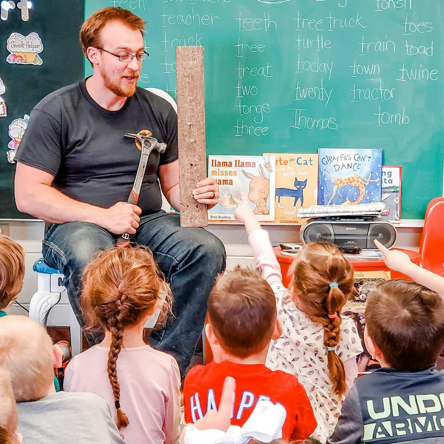 Person holding a hammer and wood plank, speaking to children in a classroom