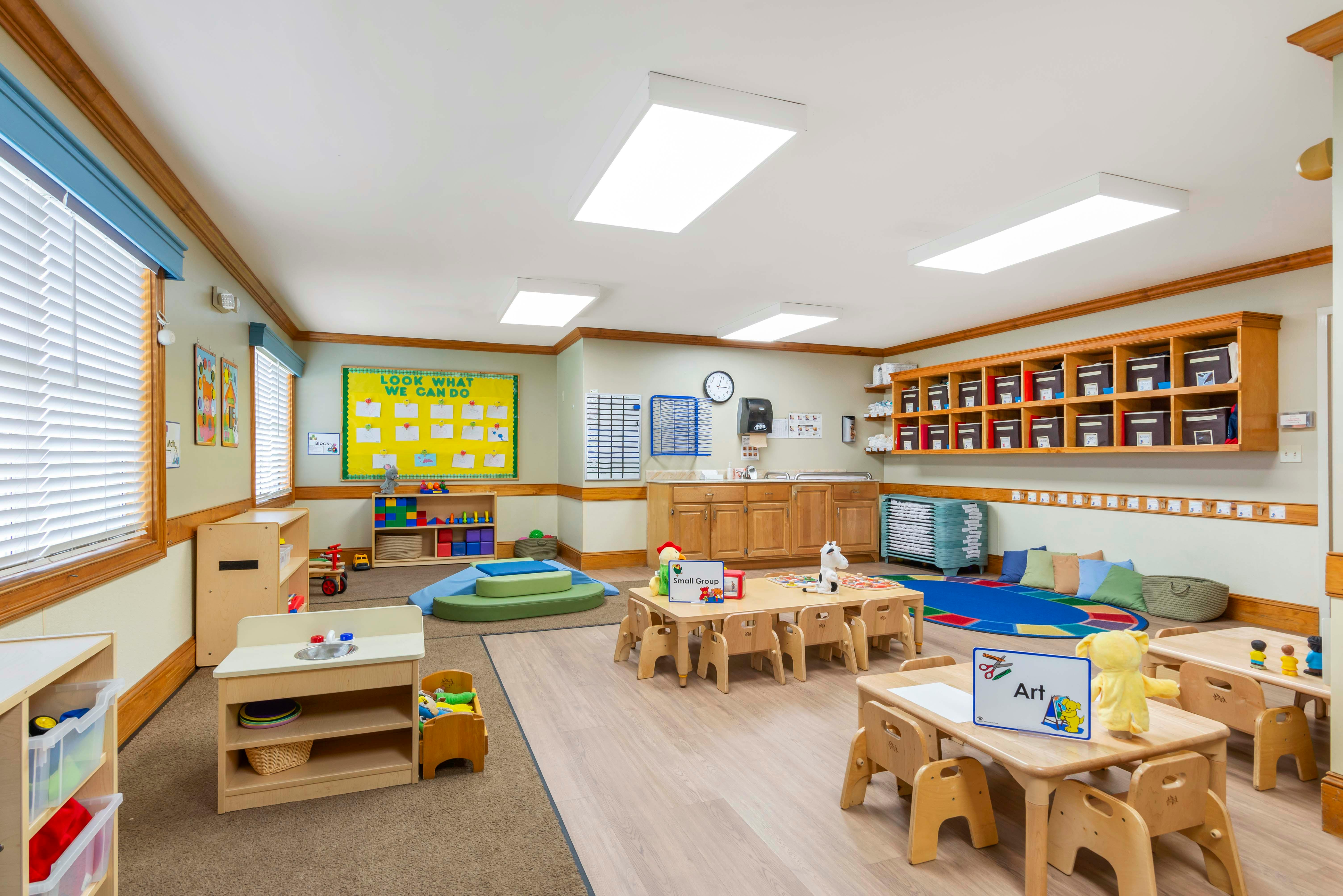 Classroom with small tables and chairs, play mats, and storage shelves