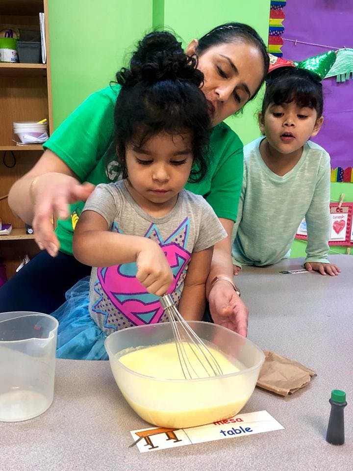 Person helping a child whisk a mixture in a bowl, another child watching