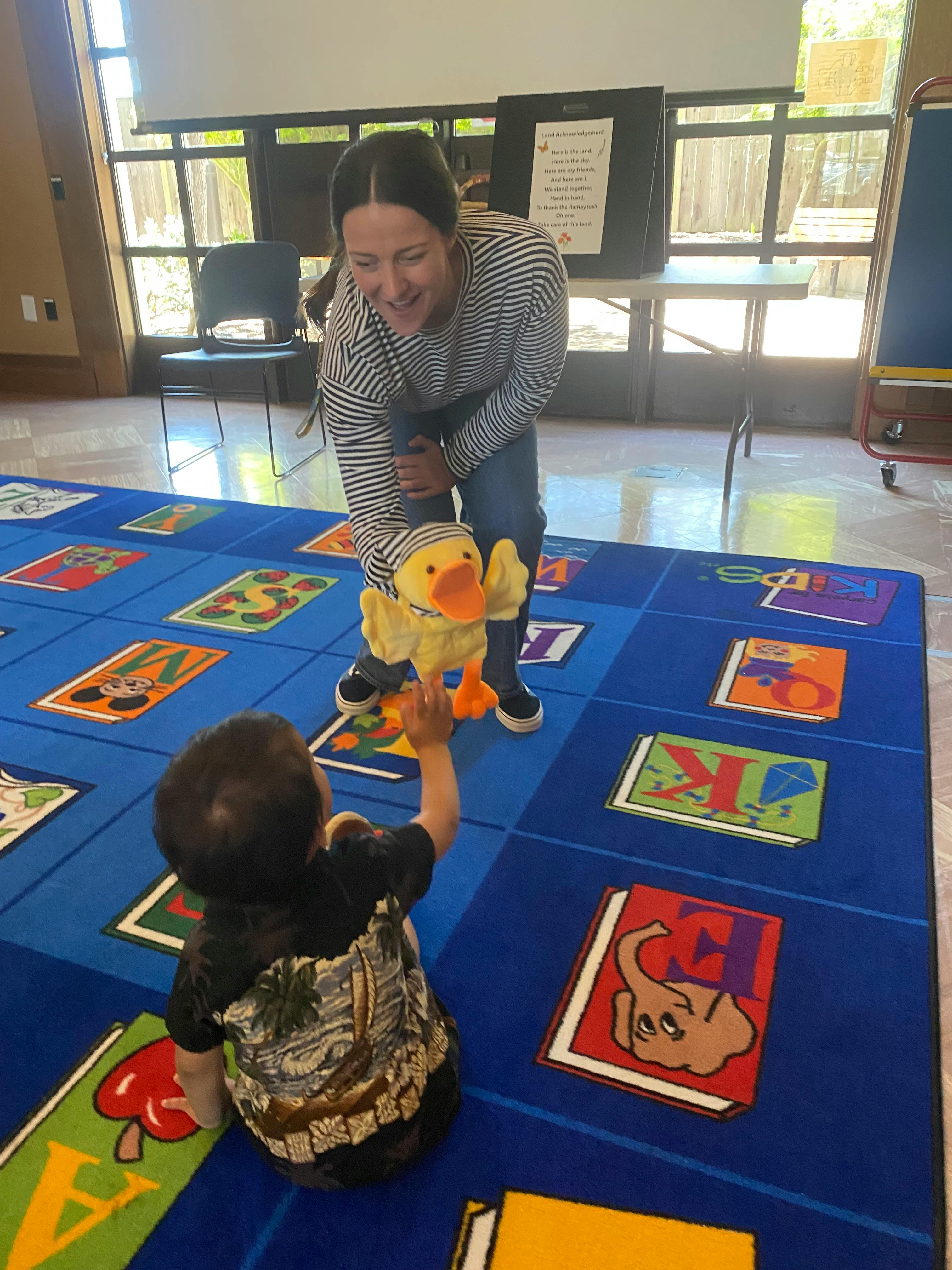 Person holding a duck puppet for a child reaching up from a blue alphabet rug