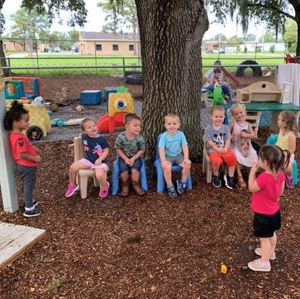 Children sitting on chairs and standing around a tree on a playground