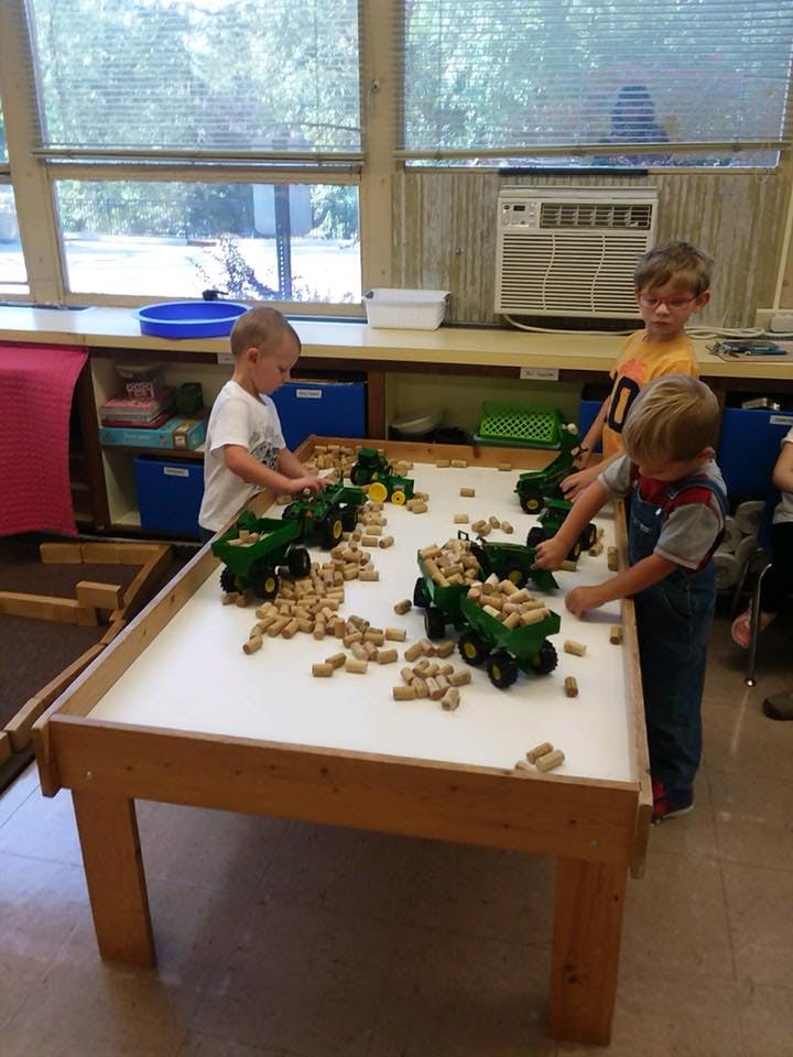Three children playing with toy tractors and small blocks on a table