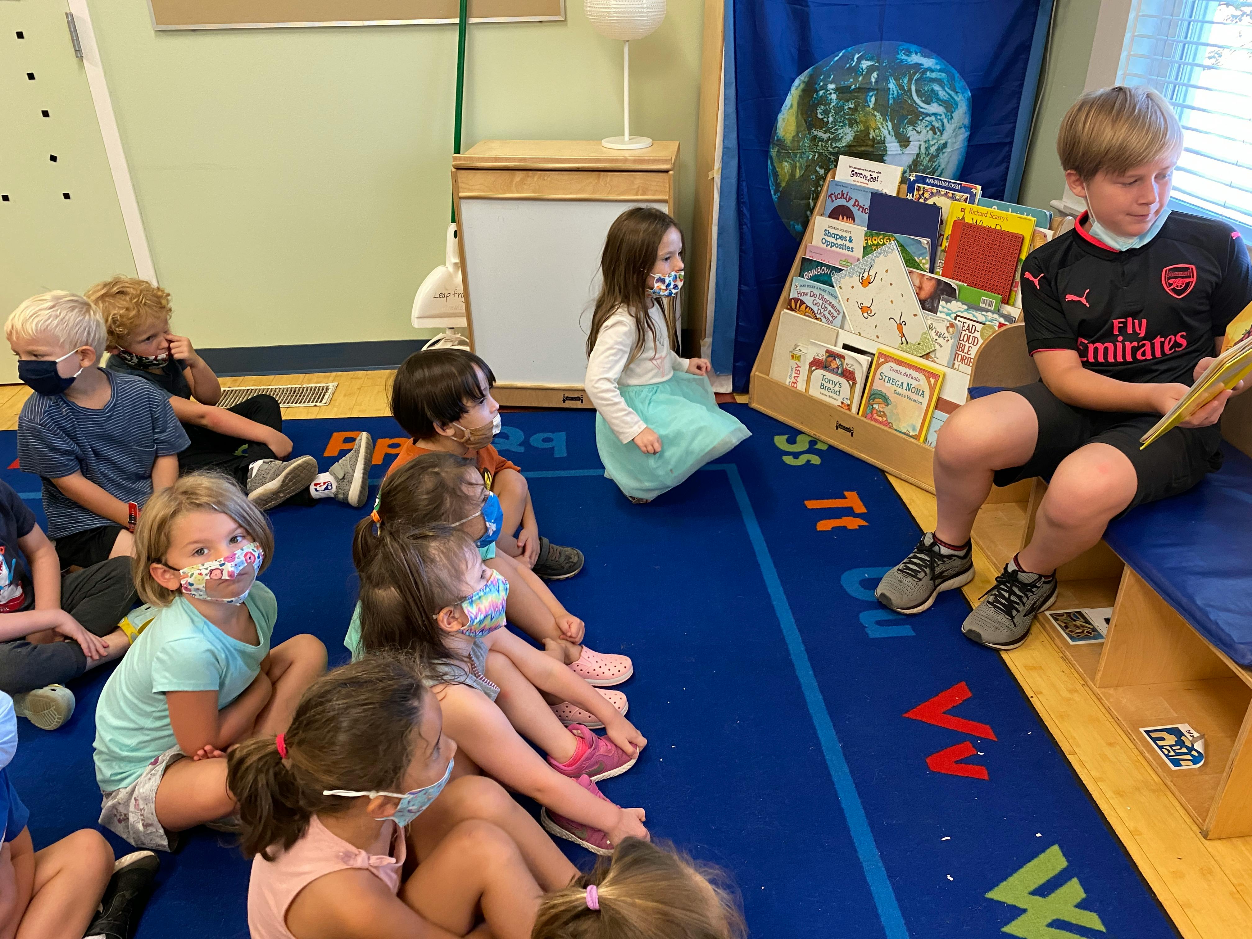 Children sitting on a rug and a teen reading a book in a classroom