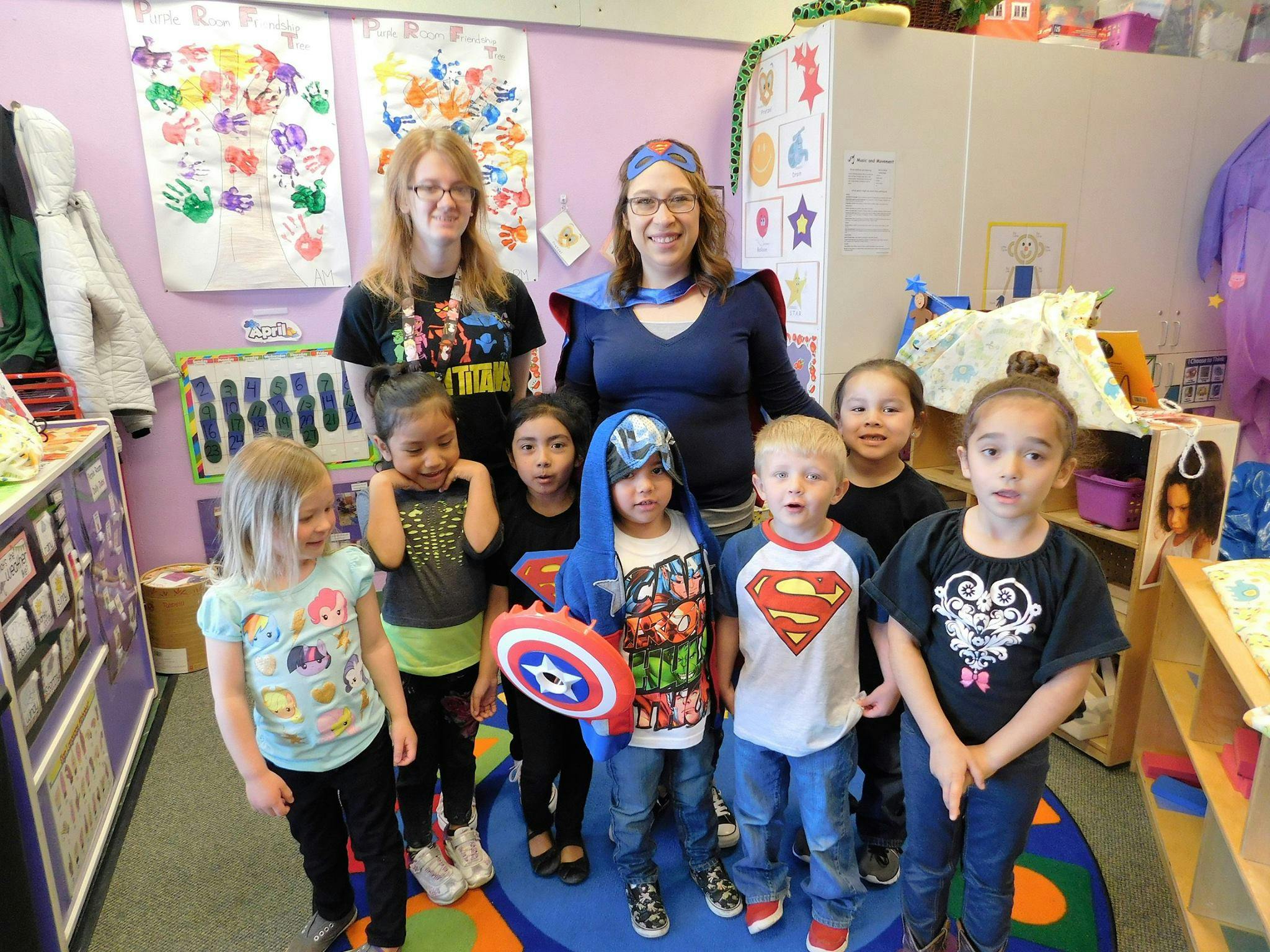 Two adults and several children in a classroom, some dressed as superheroes