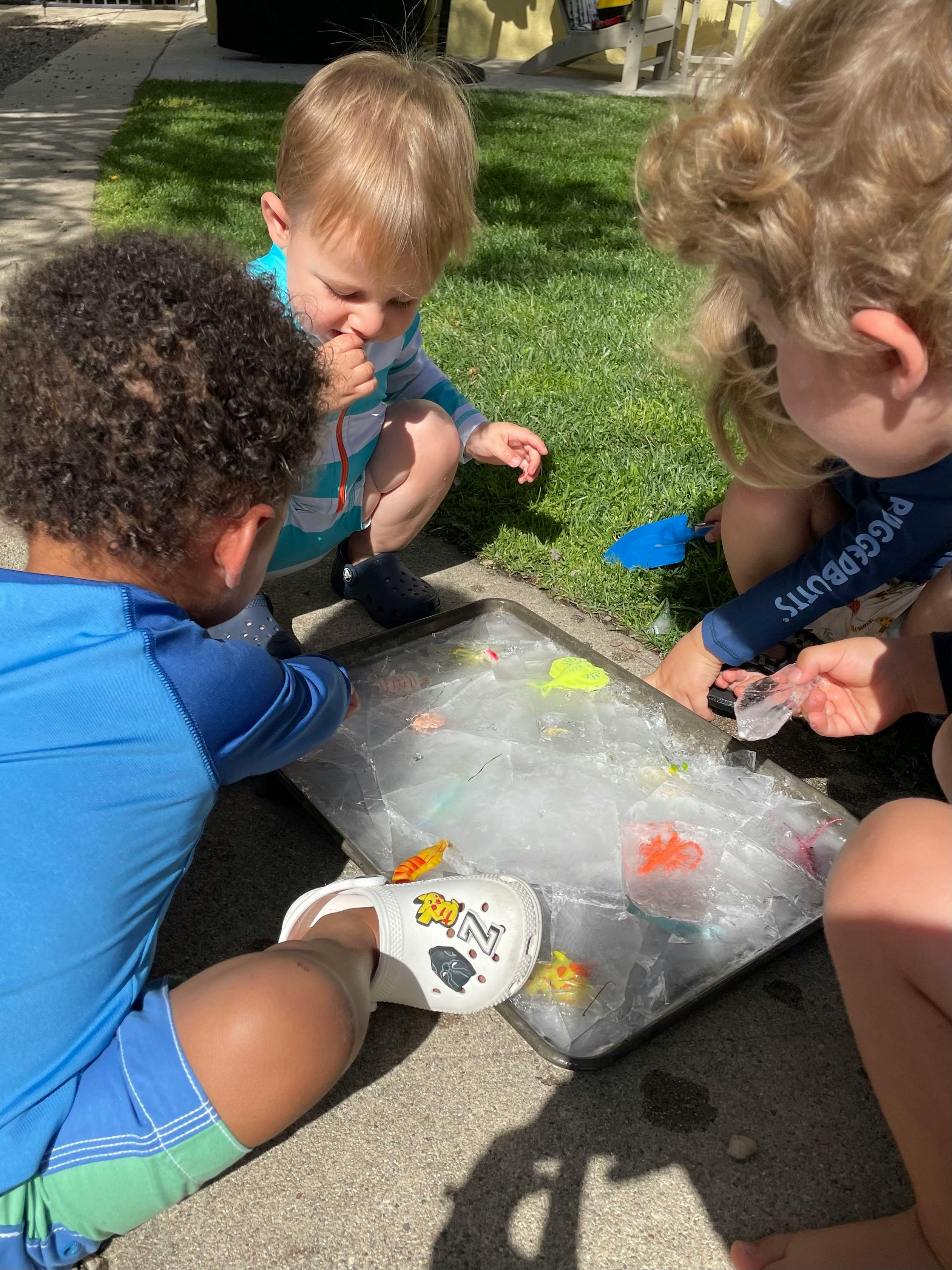 Three children playing with ice and small toys in a tray outdoors