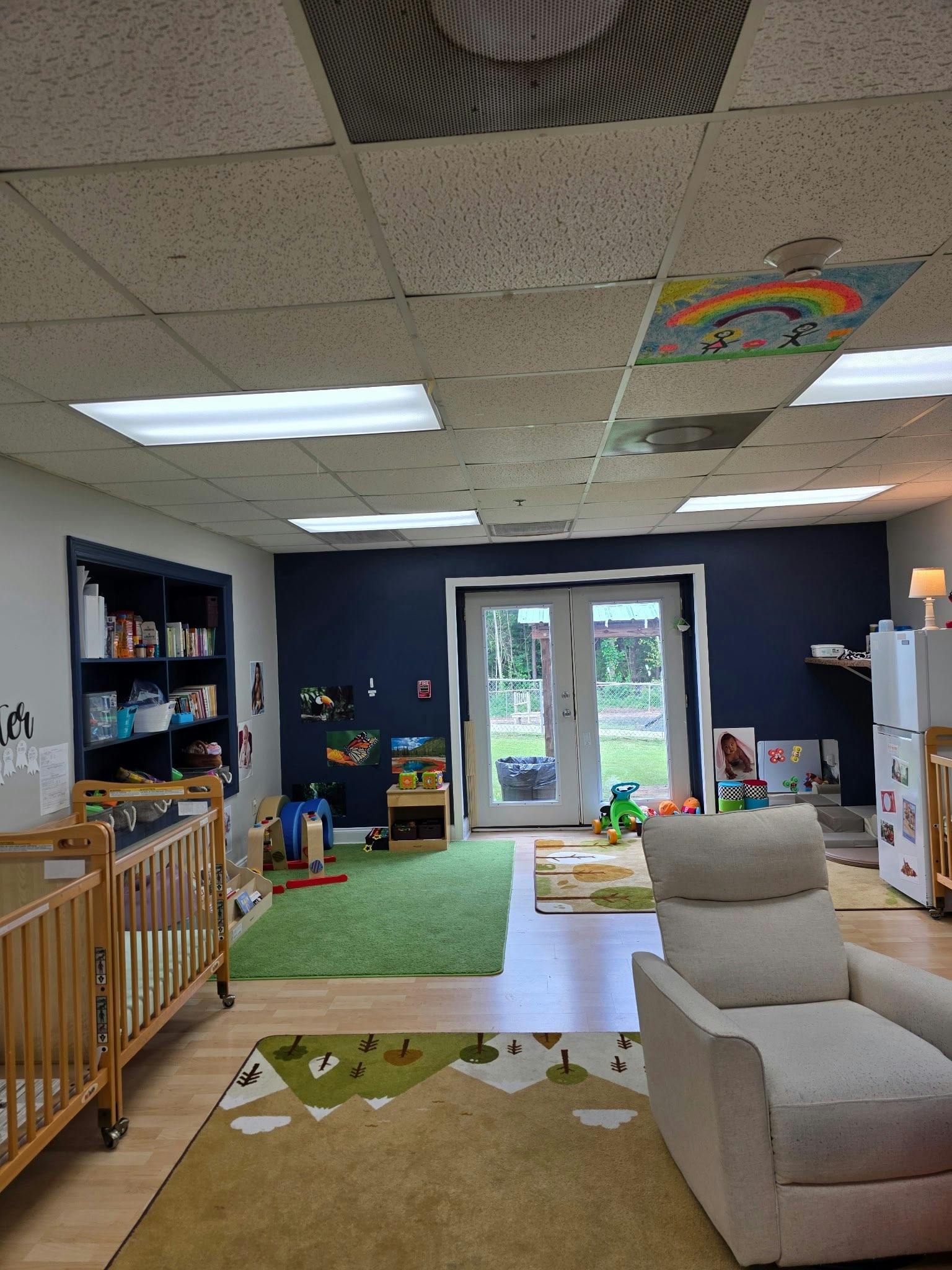 An indoor play area with cribs, a recliner, and colorful rugs on the floor