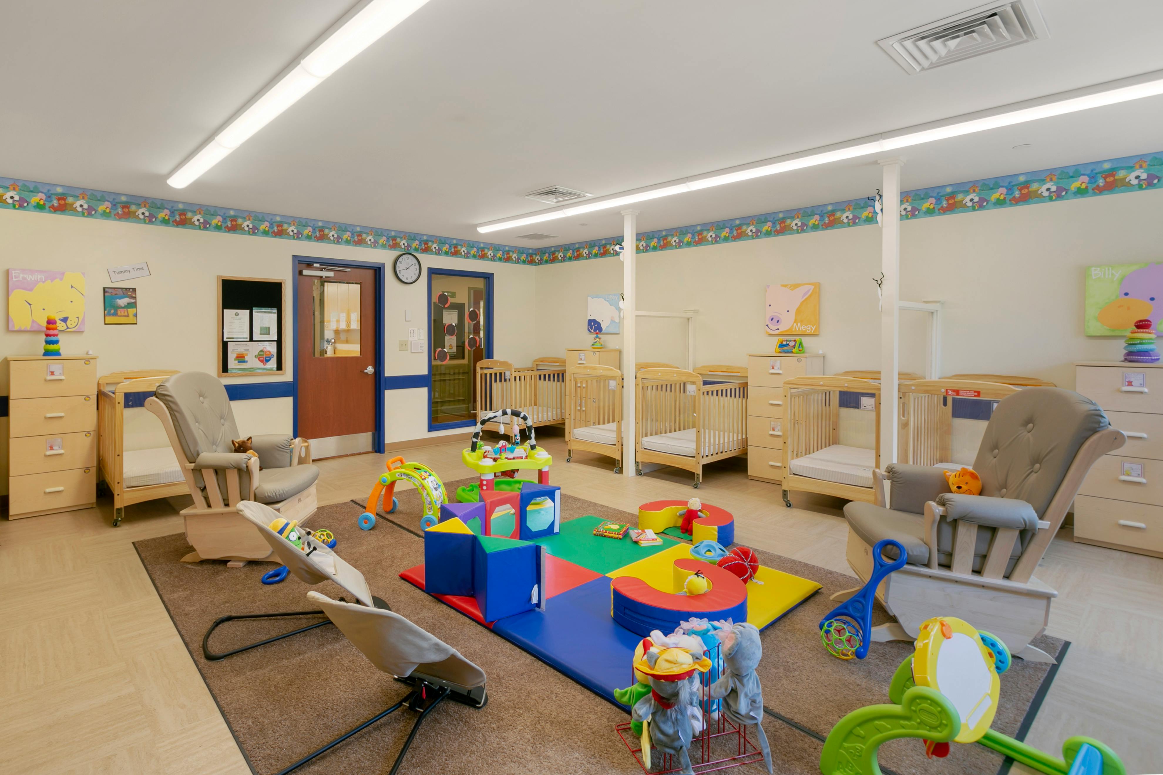 A colorful childcare room with cribs, rocking chairs, and playmats with toys