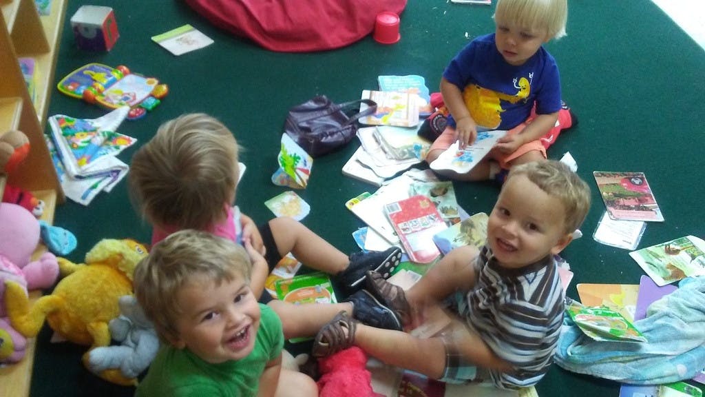 Multiple children sitting on a floor surrounded by books and toys
