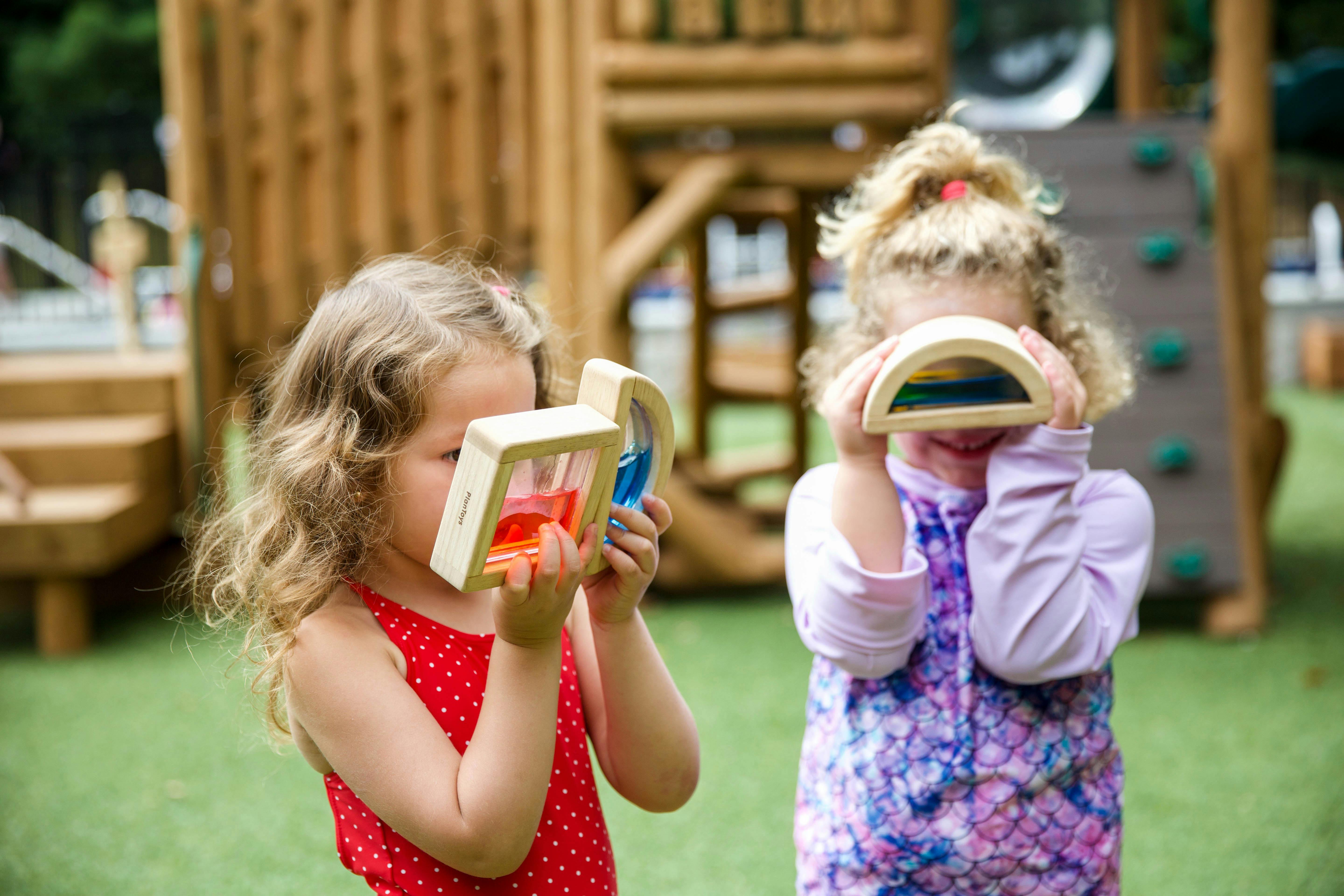Two children holding up wooden toys to their faces on a playground