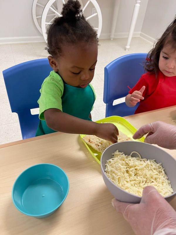 Child at a table reaching for food on a tray, person in gloves holding a bowl