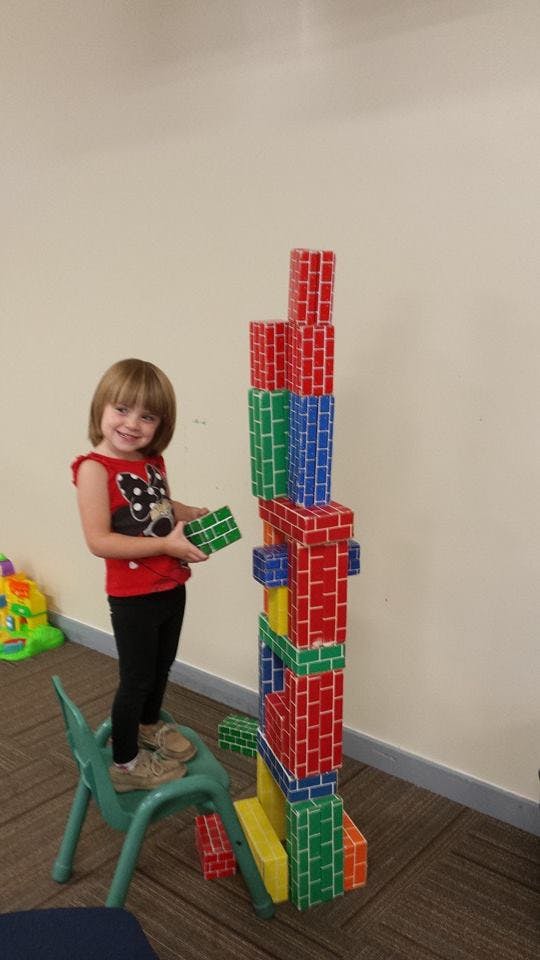 Child standing on a chair, holding a block next to a tall block tower