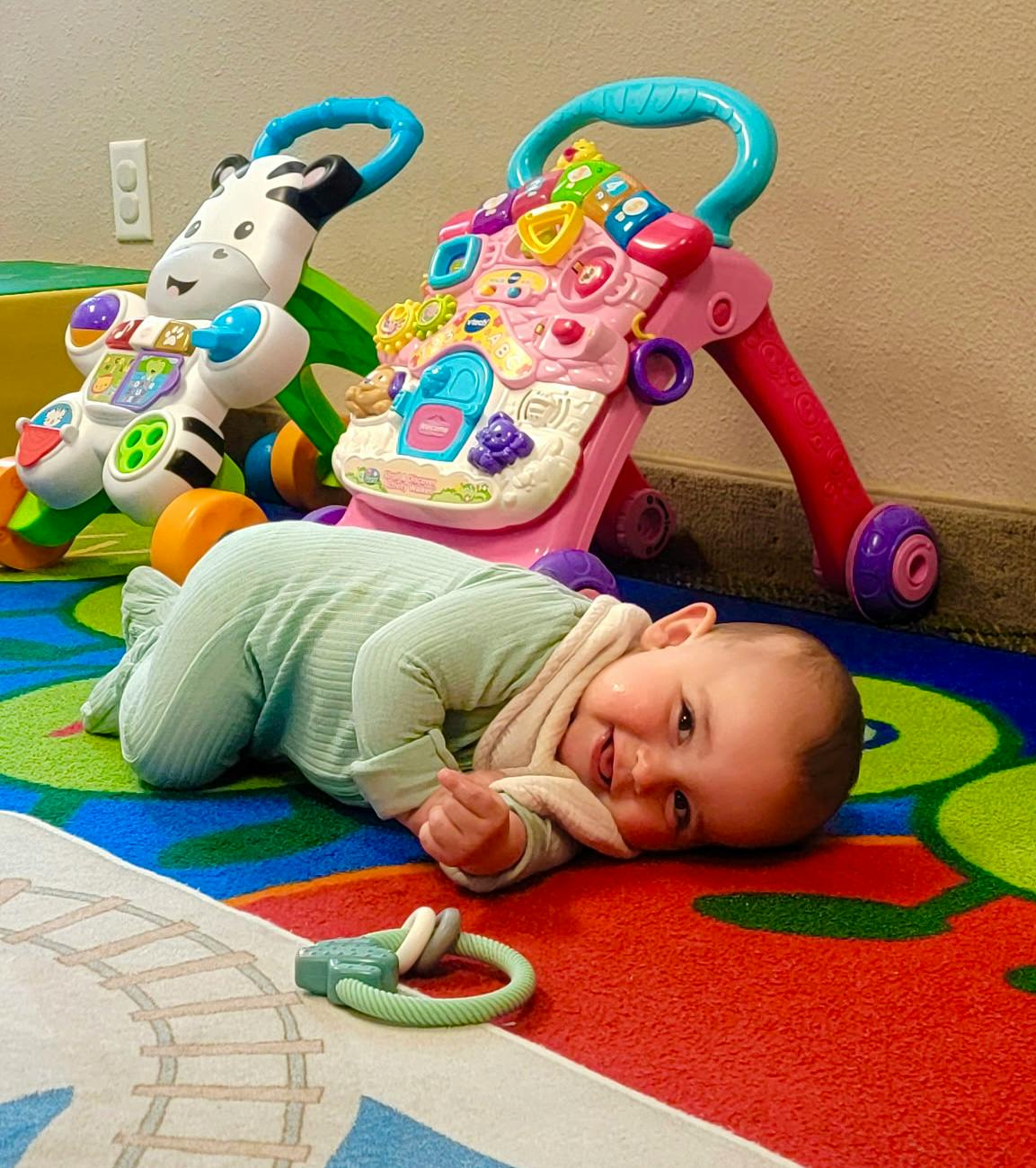A child lying on a colorful patterned floor near toy walkers