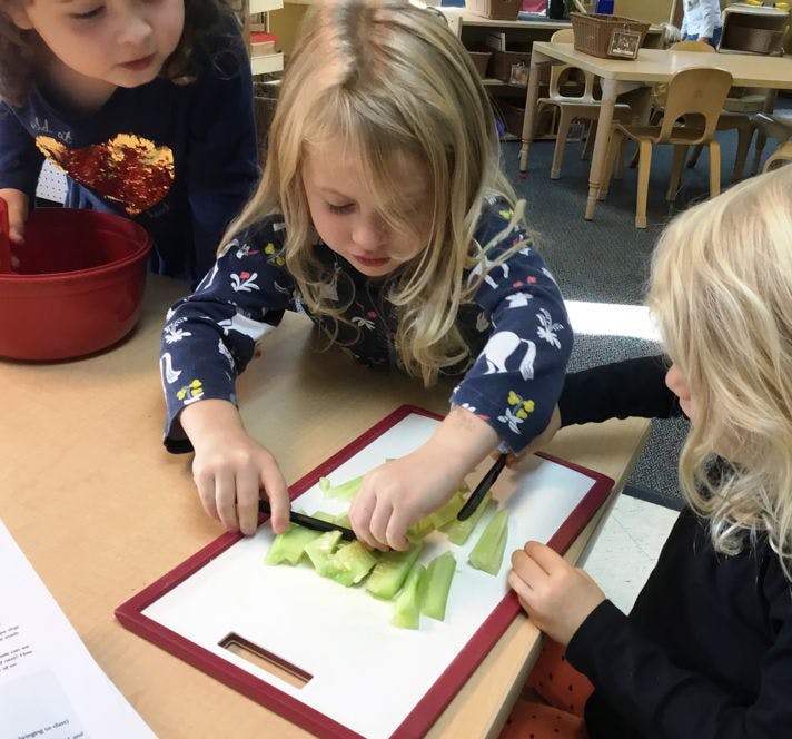 Child cutting celery on a white cutting board with another child watching