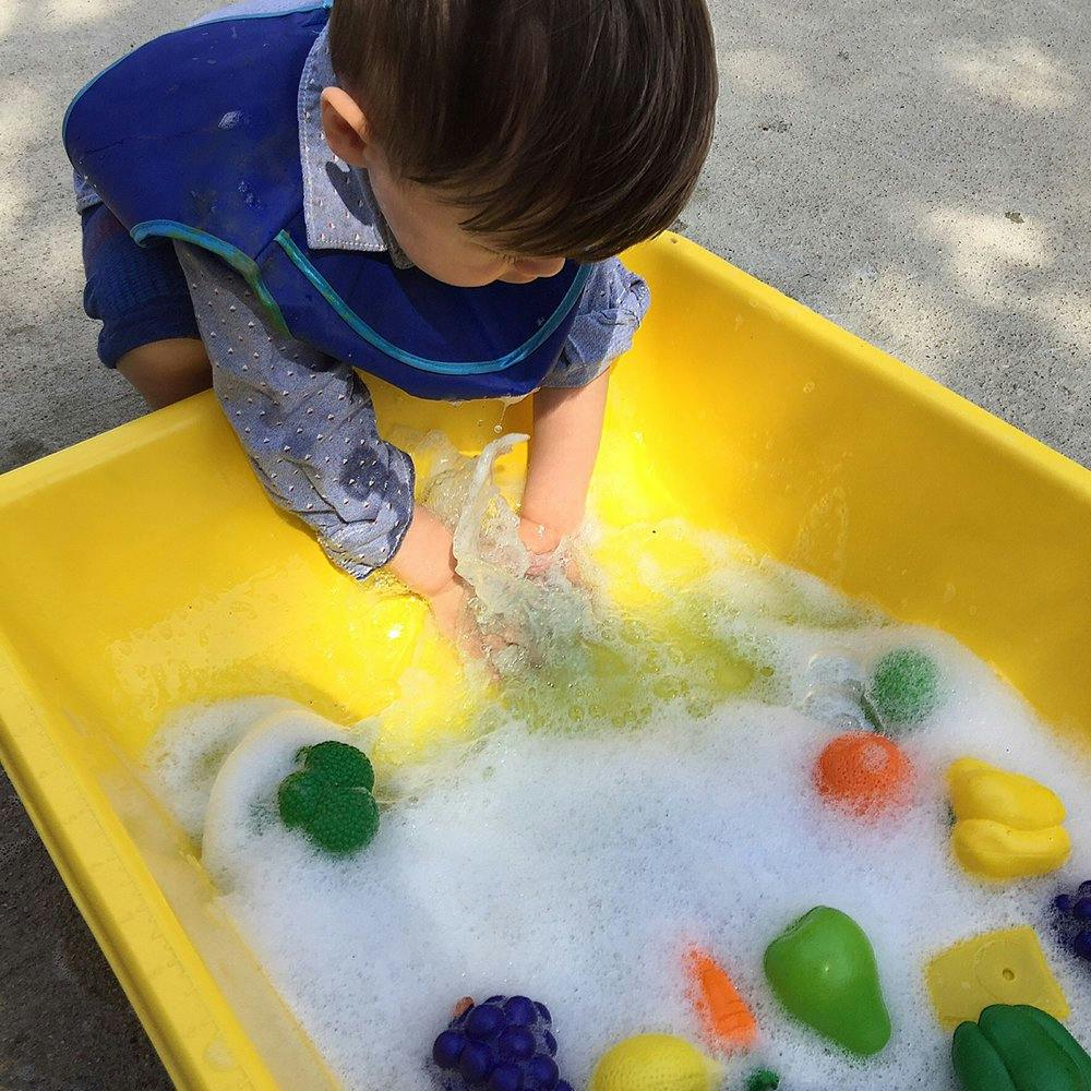 Child playing with water and toy vegetables in a yellow bin
