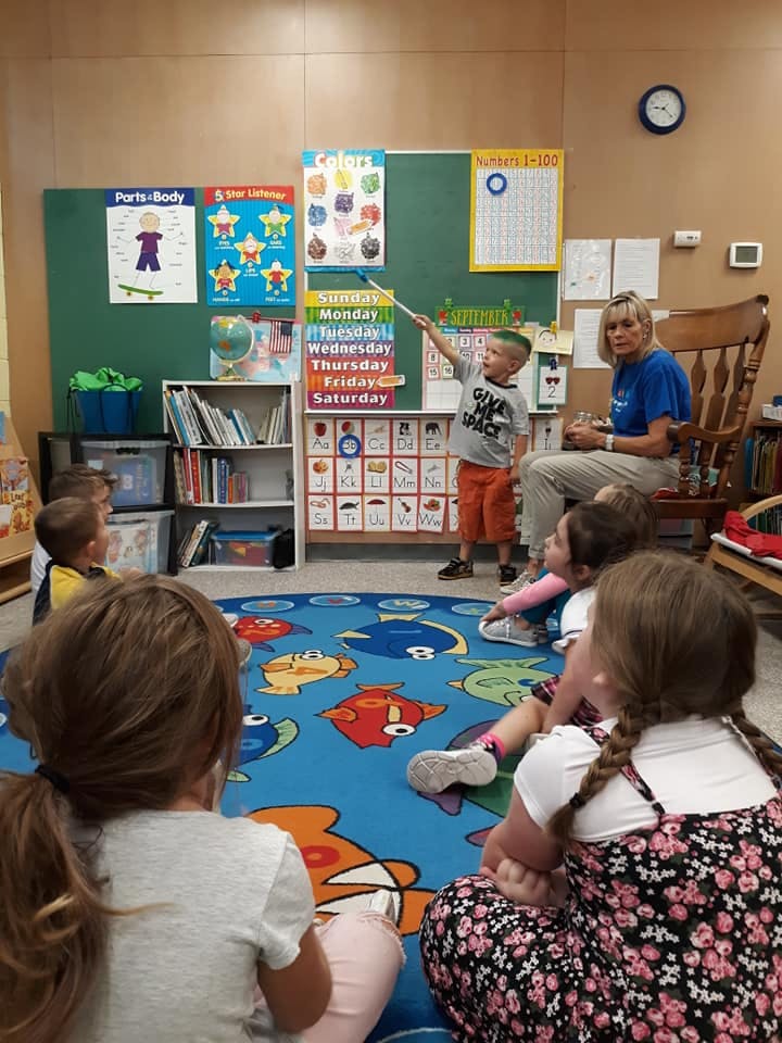 Child pointing at a calendar in a classroom with an adult and other children