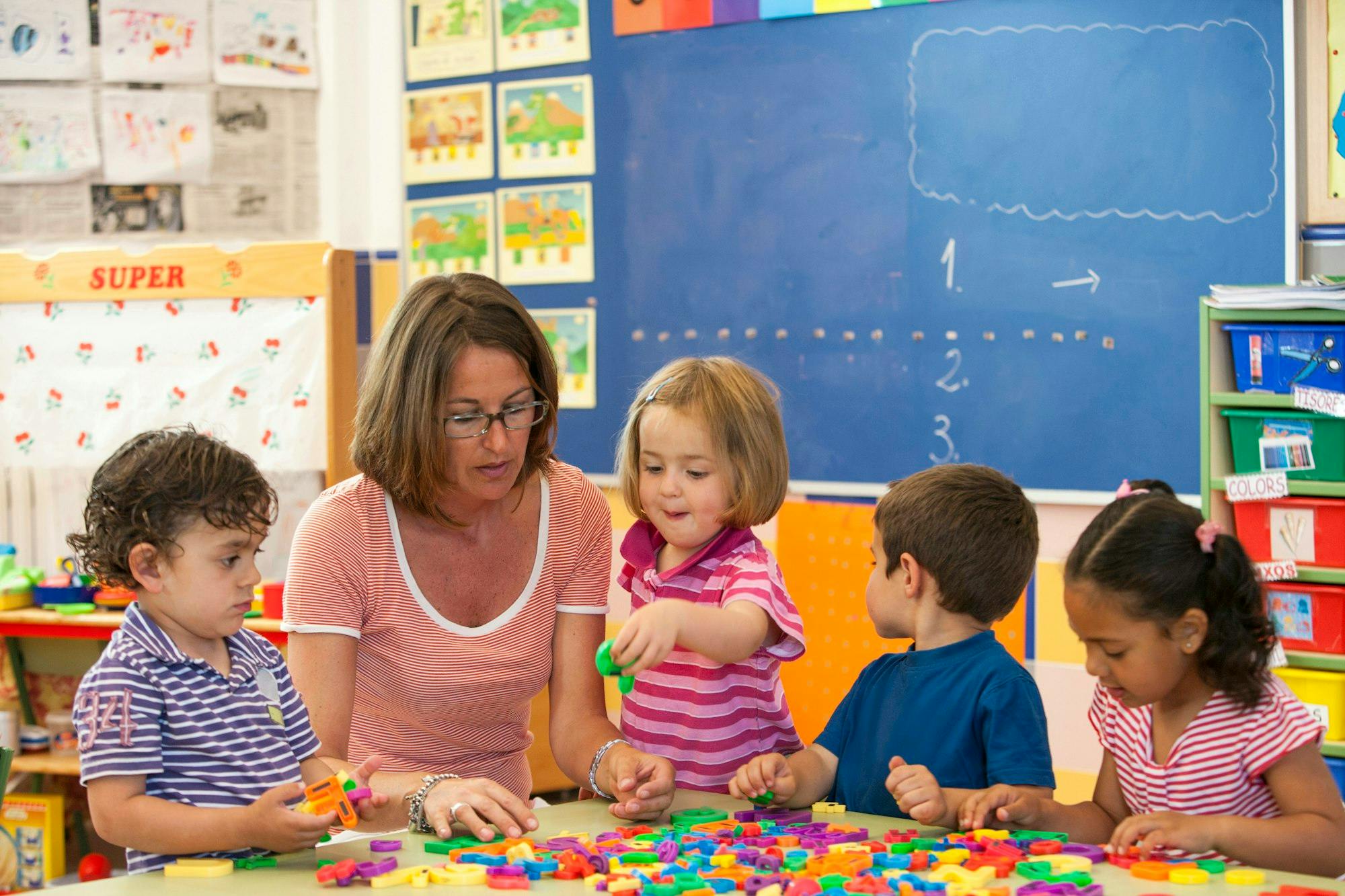 Adult and three children playing with colorful alphabet letters at a table in a classroom