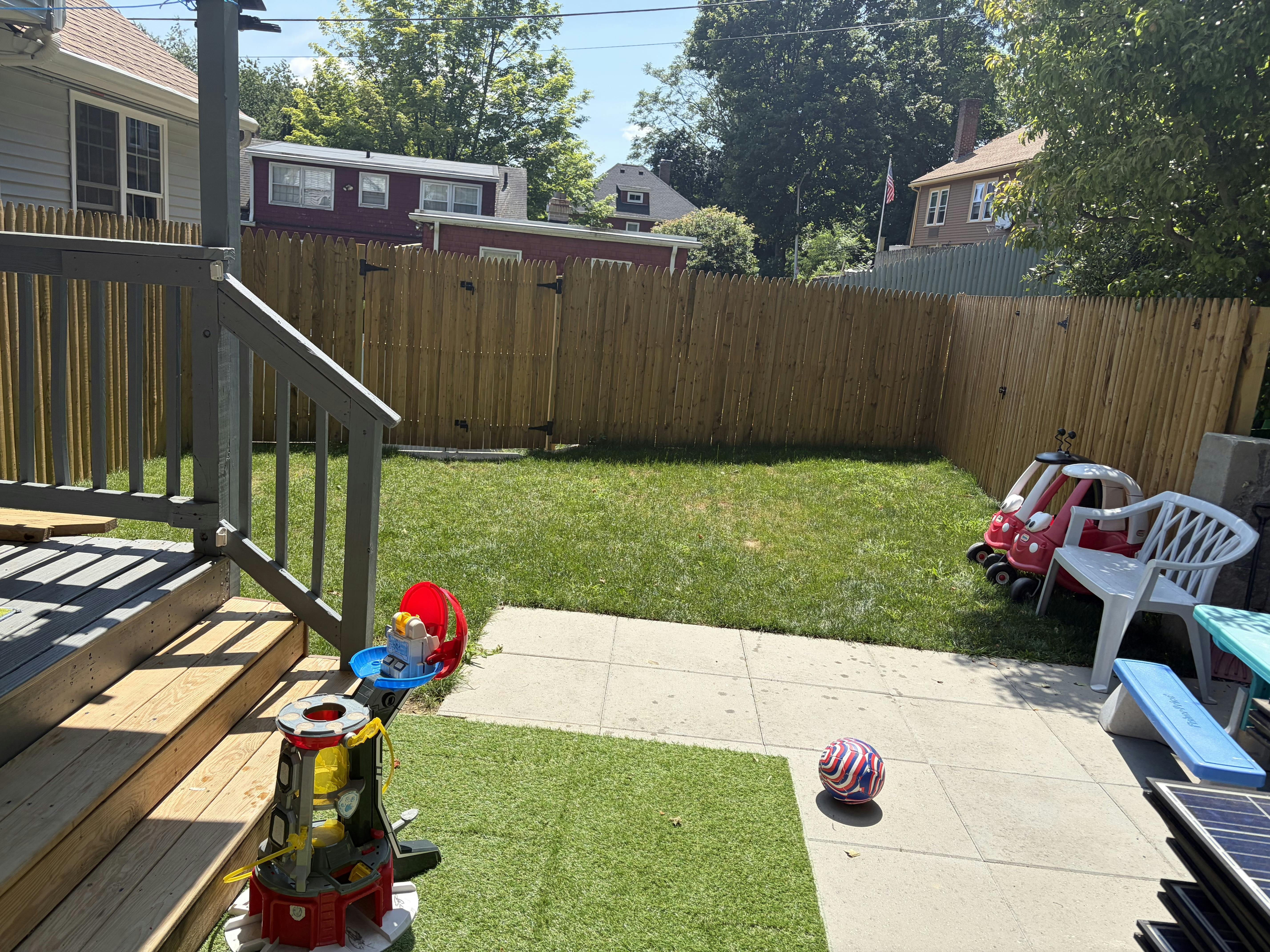 Backyard with patio, lawn, wooden fence, and scattered child toys