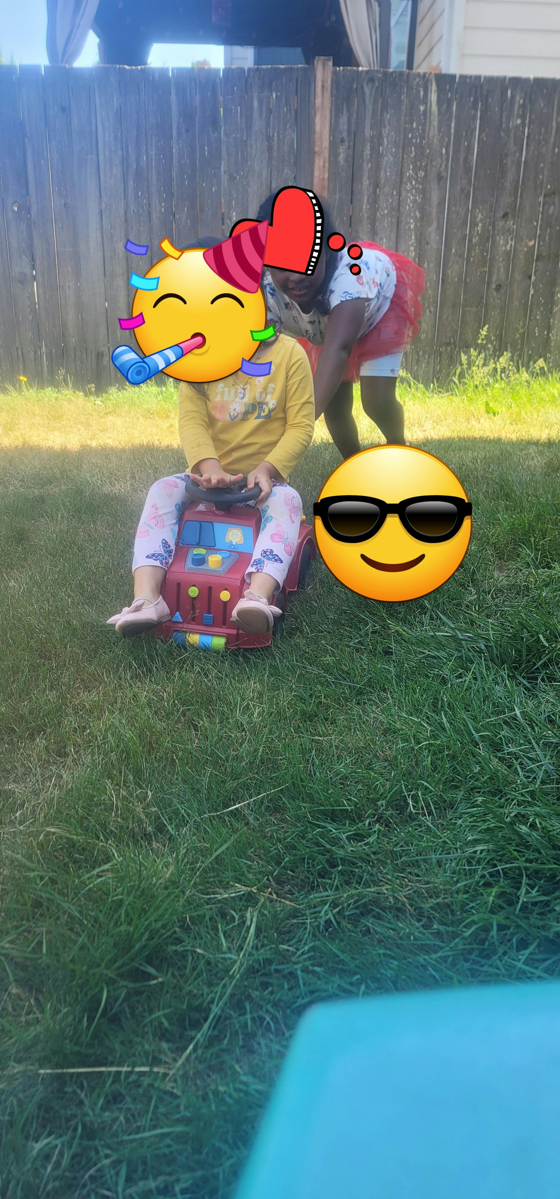 Child sitting in a toy car on grass with another person standing behind