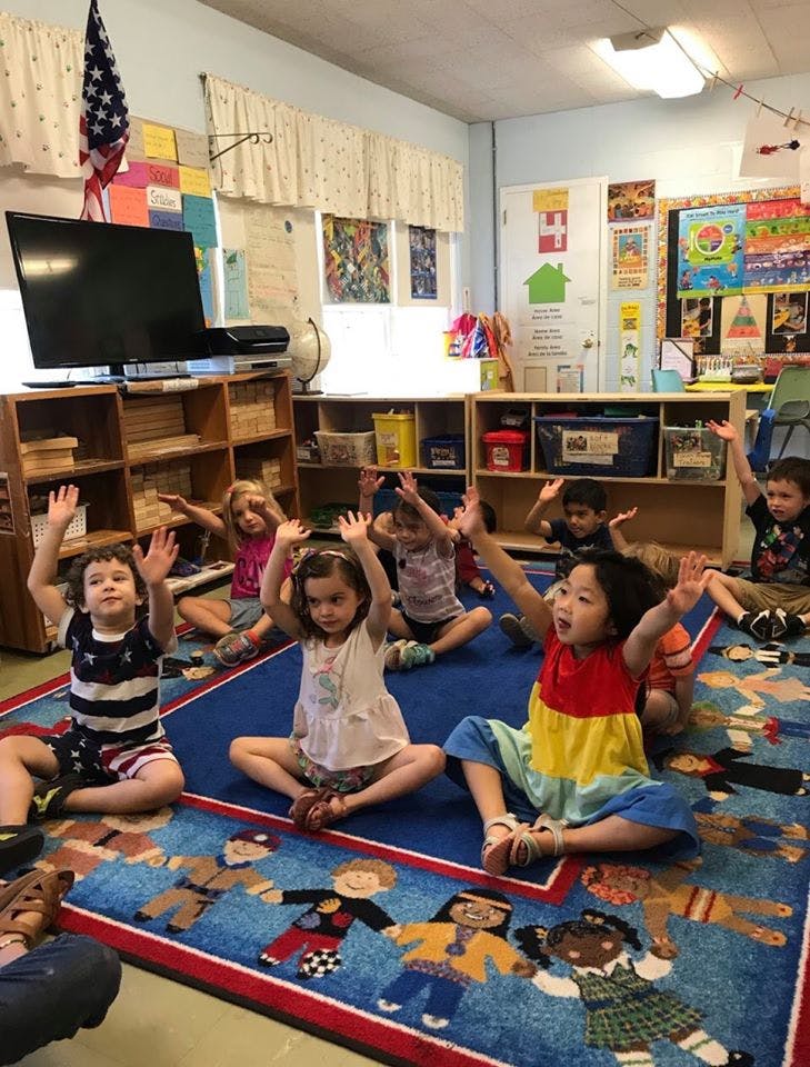 Children sitting on a rug with their hands raised in a classroom