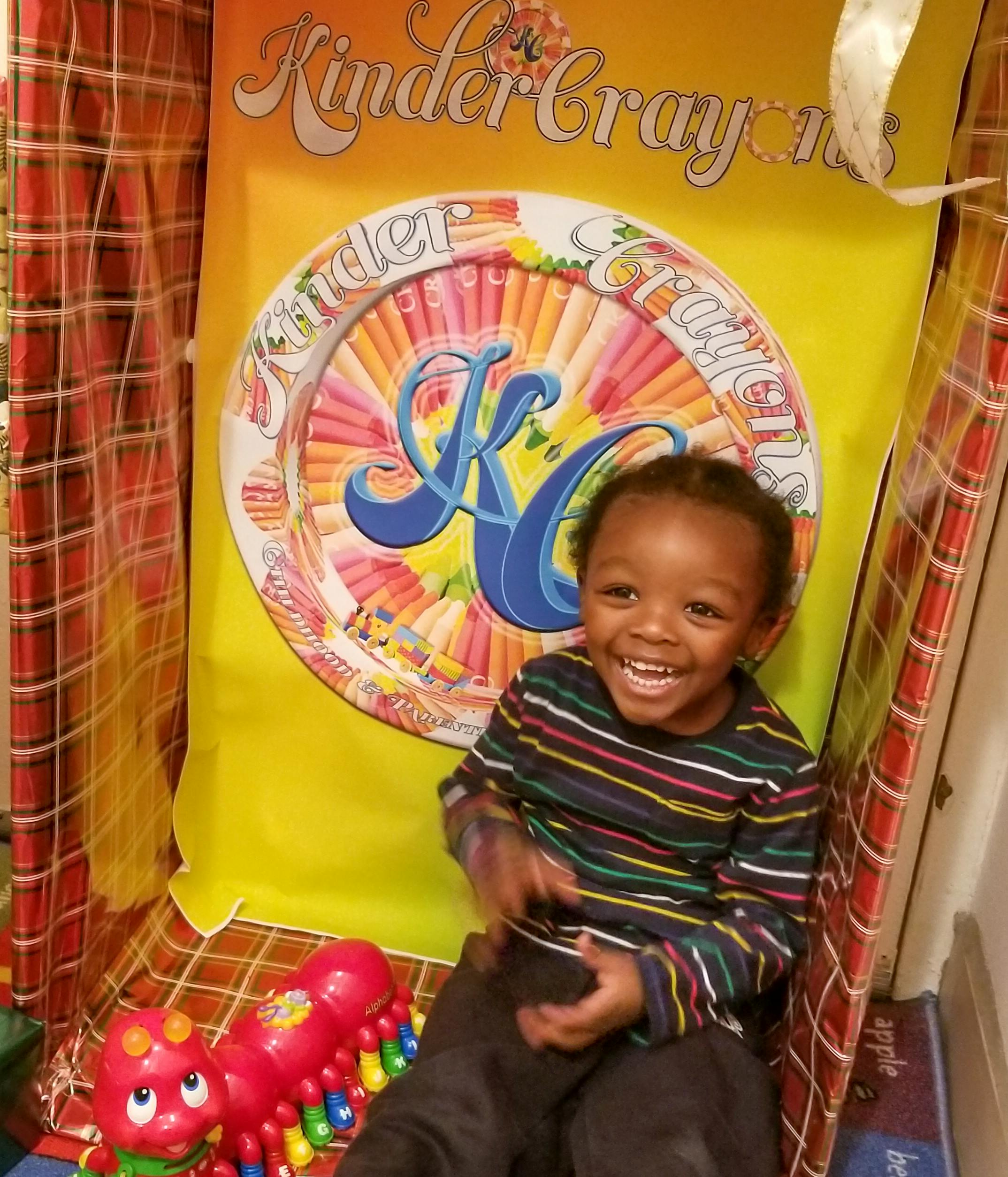 Child sitting in front of a yellow banner with "Kinder Crayons" text