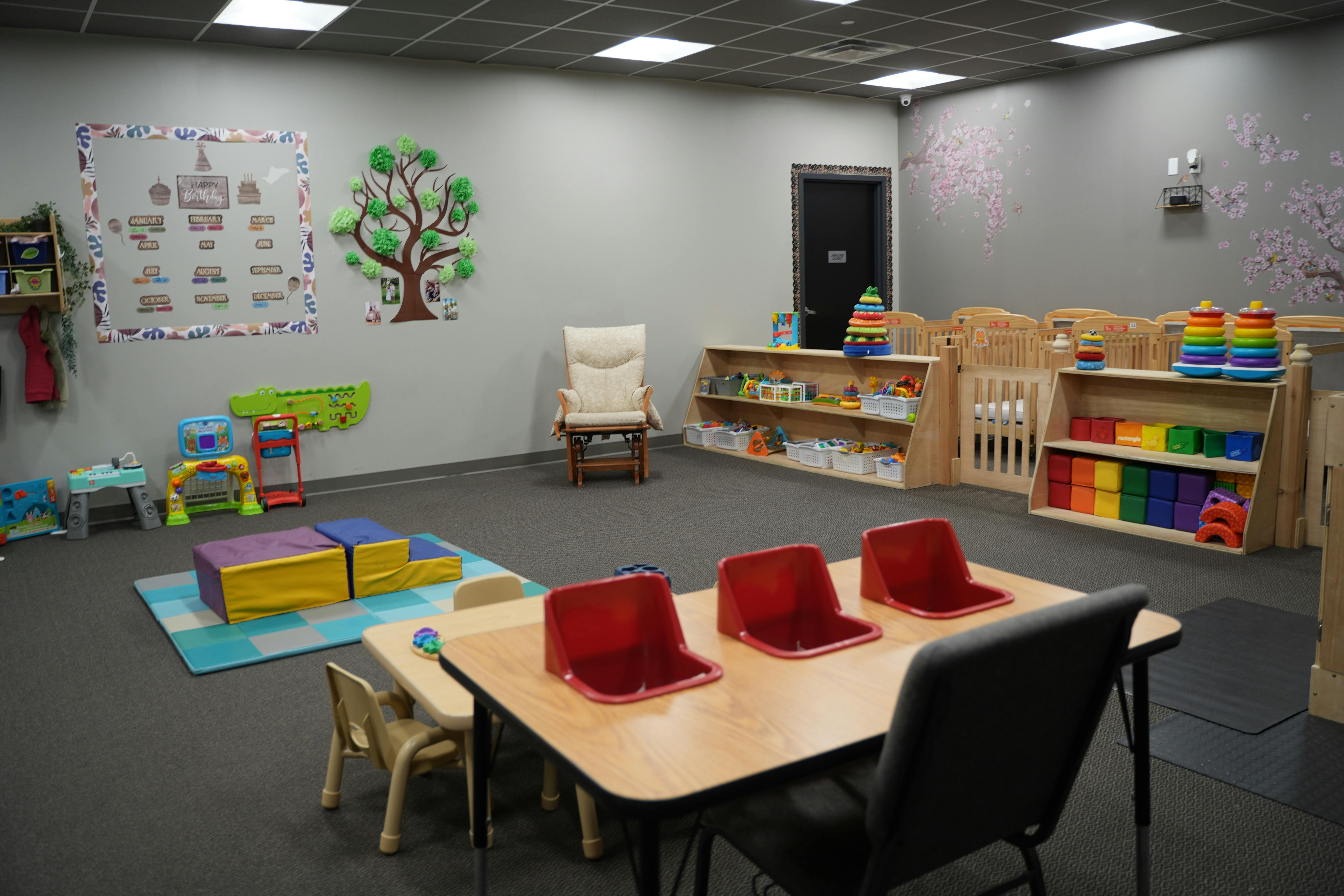 Playroom with cribs, small tables and chairs, and a birthday board on the wall