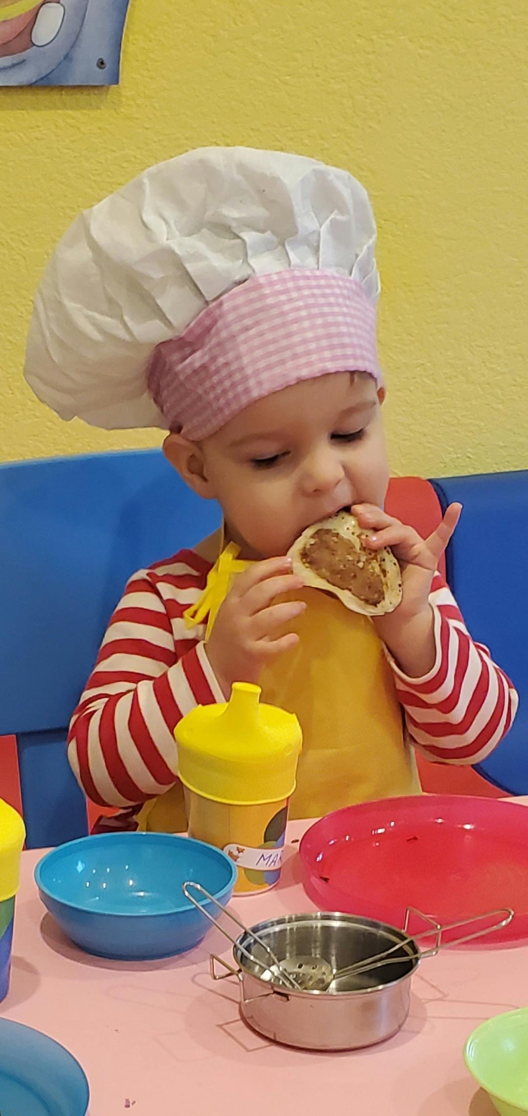Child wearing a chef hat and apron eating a toy taco at a play table