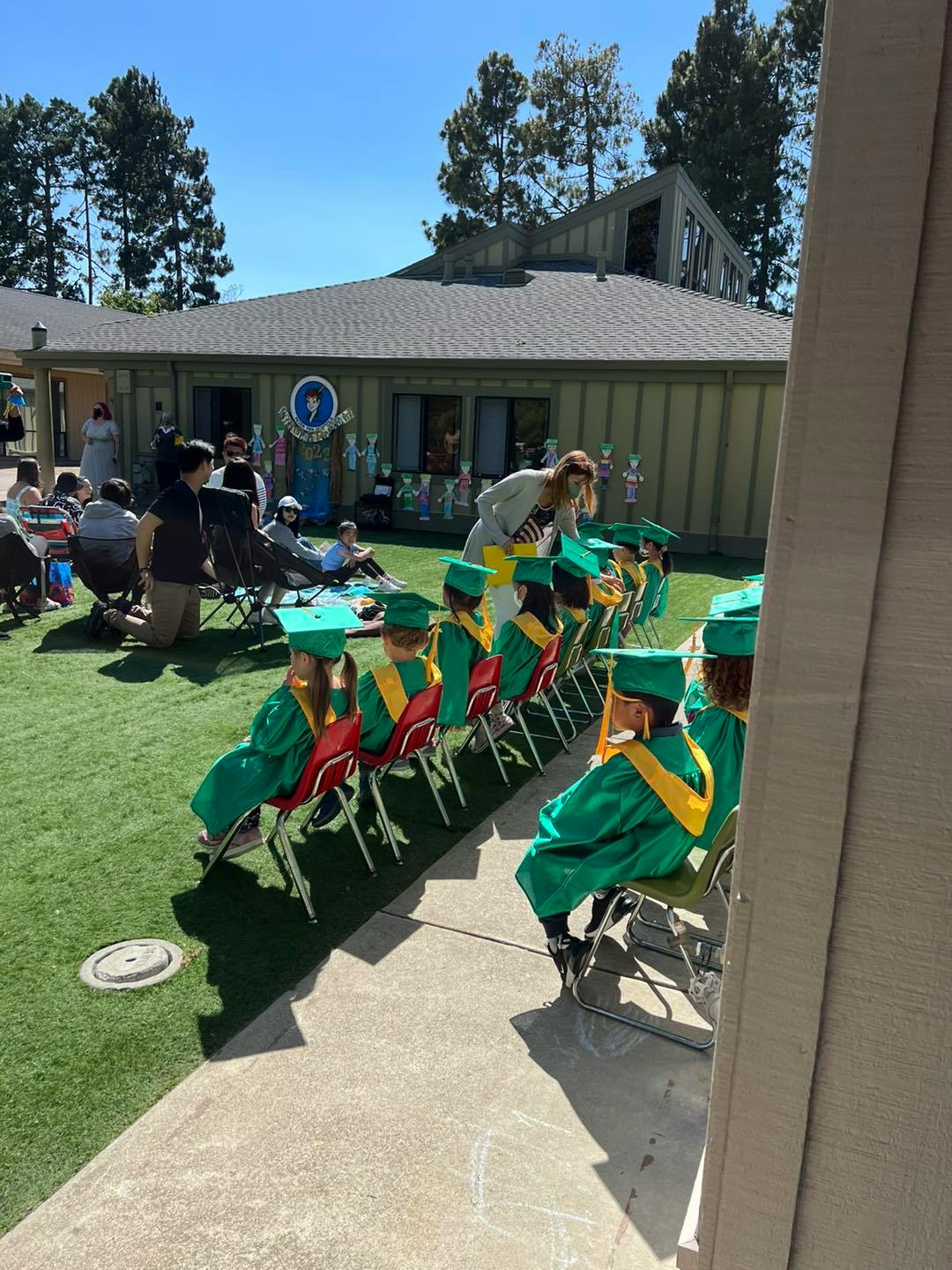 Children in green graduation gowns and caps sitting in chairs outdoors