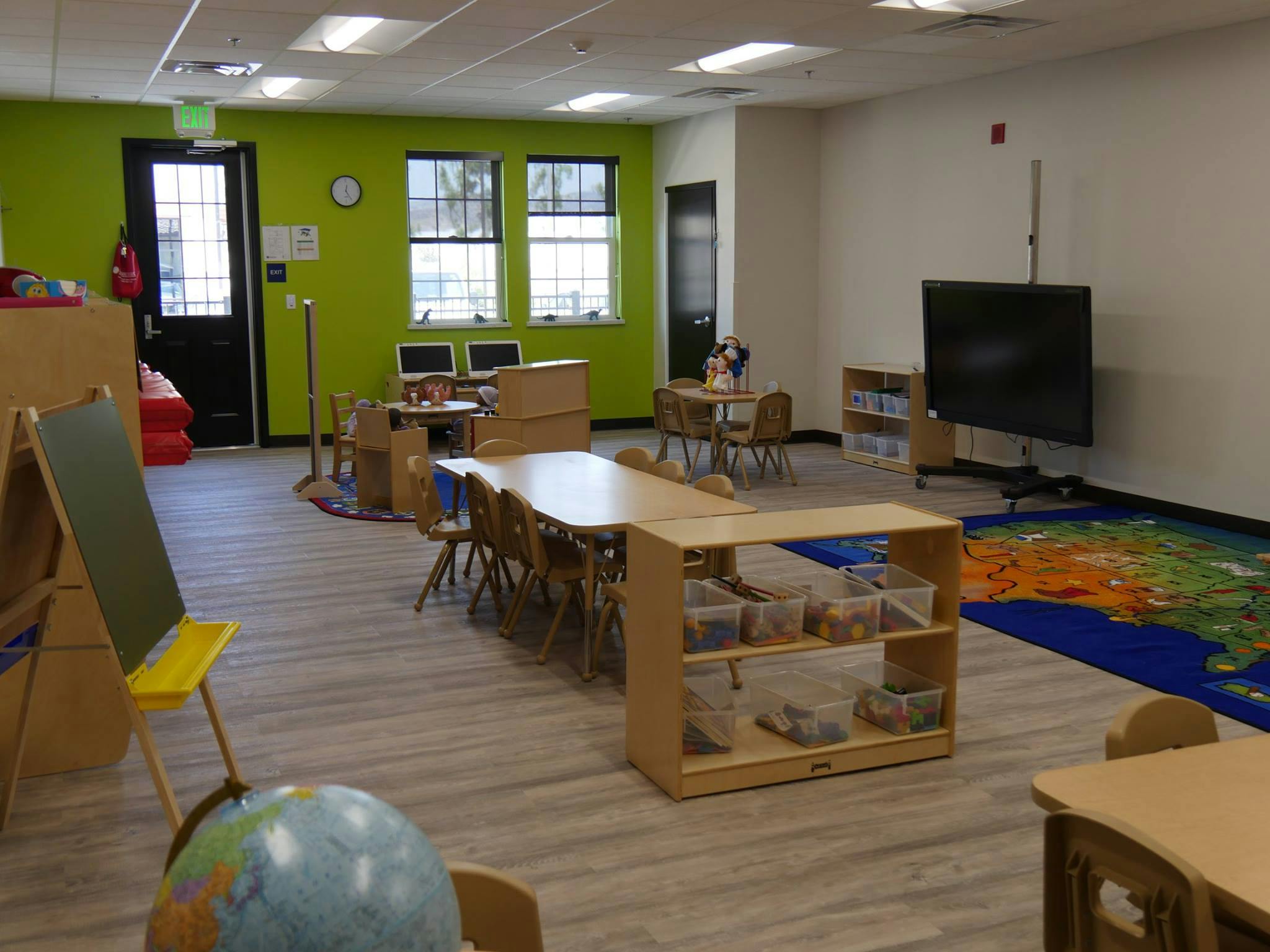 Empty classroom with small tables and chairs, a large screen, and a world map rug