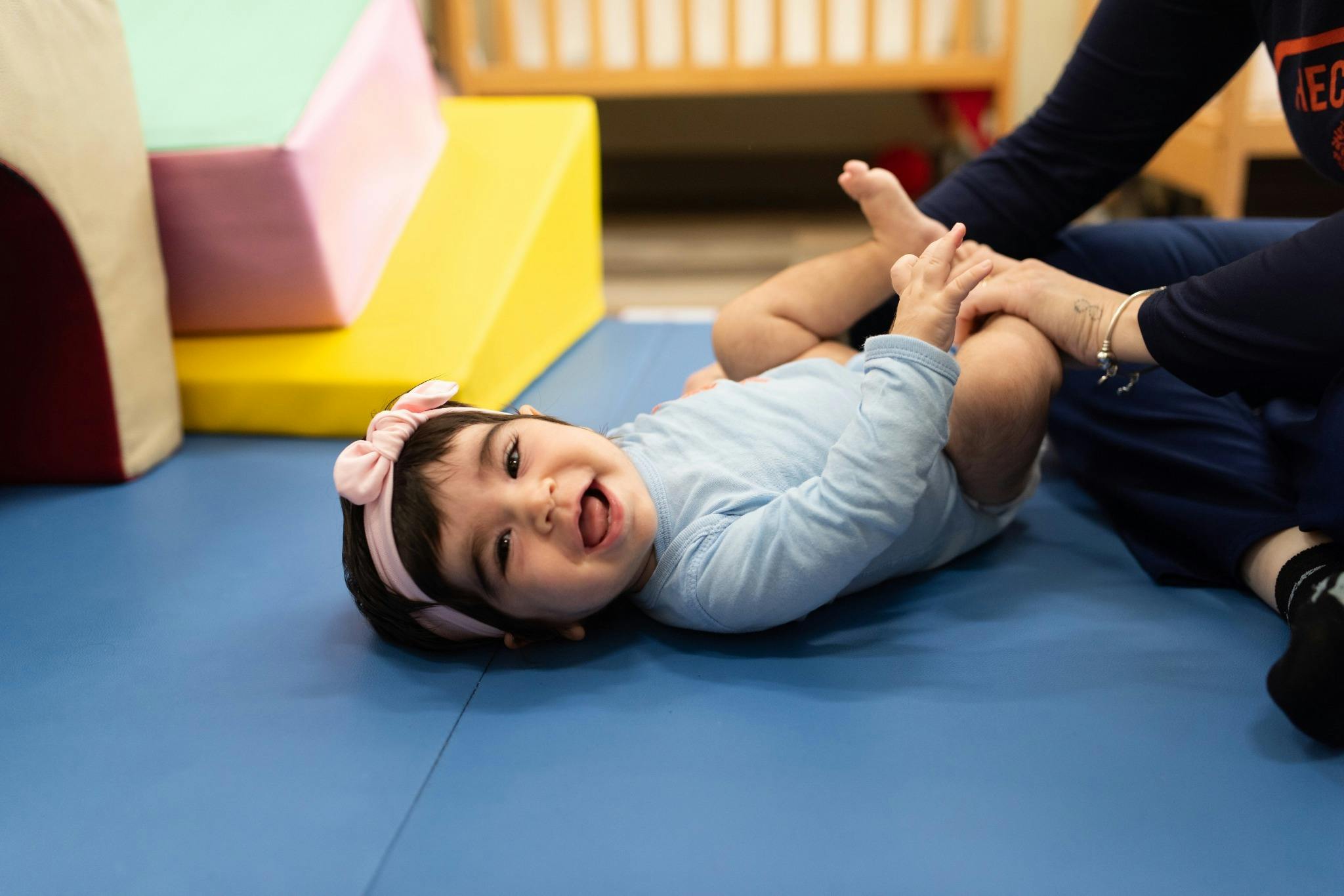Child lying on a blue mat, looking up, while a person holds their feet
