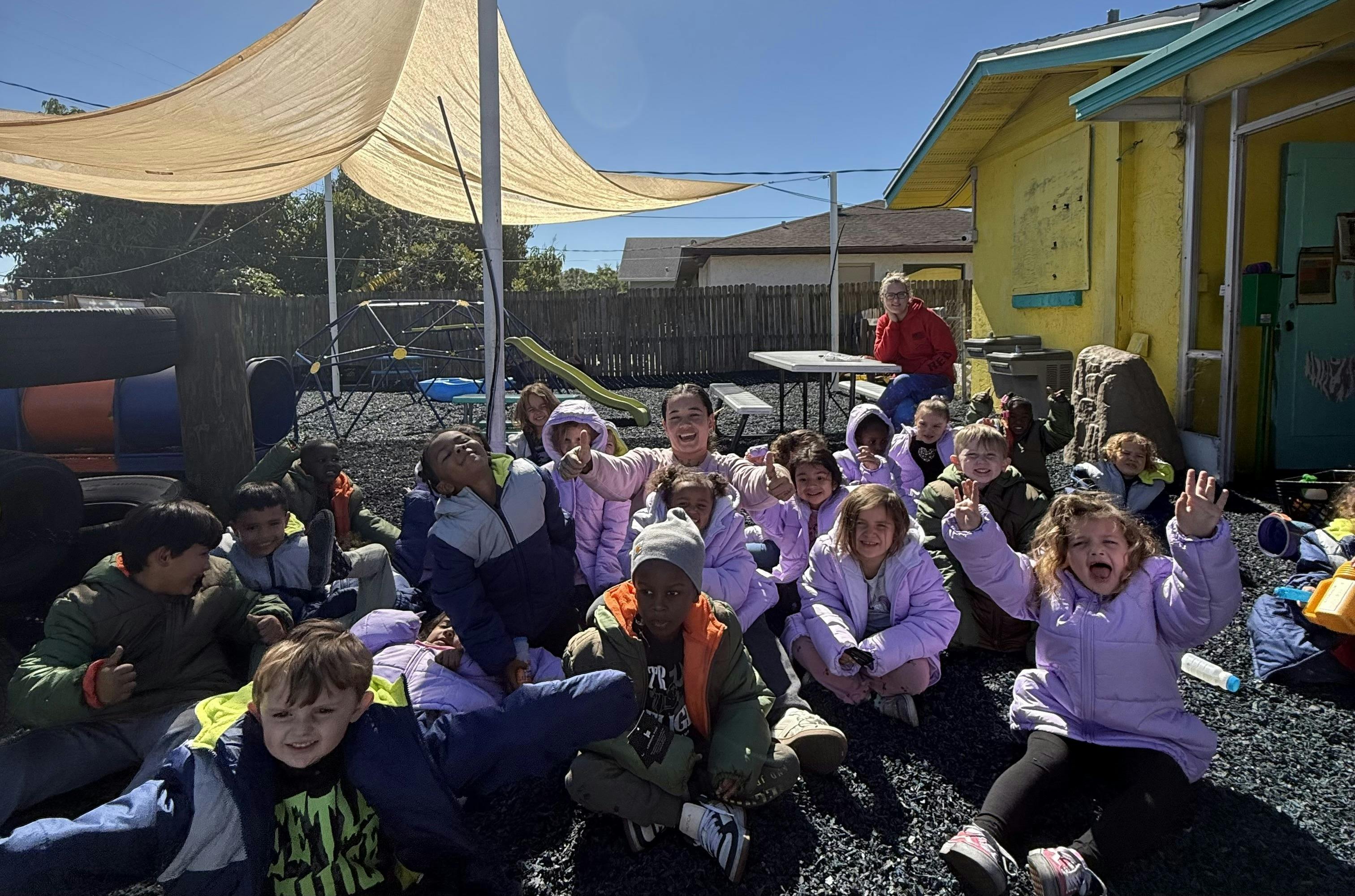 Children and a woman sitting on the ground outdoors, some looking up and smiling