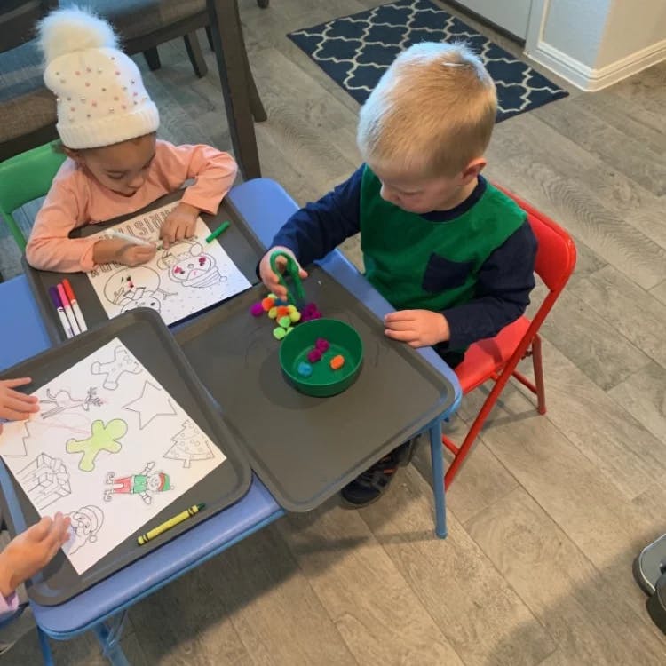 Two children at a table, one coloring and one playing with small objects