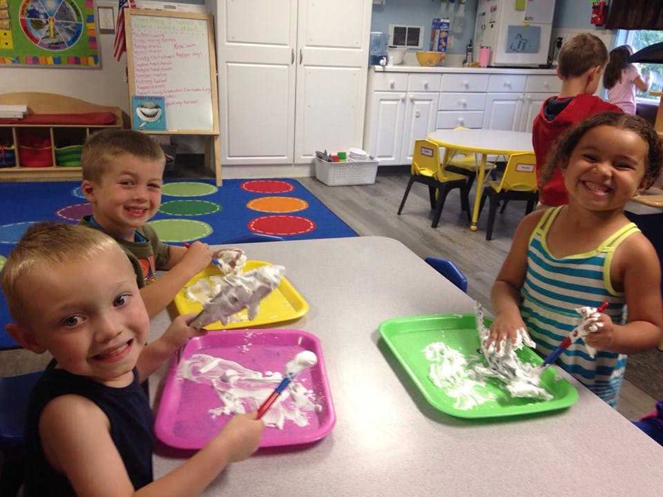 Three children playing with shaving cream on trays in a classroom