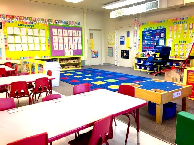 Empty classroom with small tables and chairs, colorful rug, and bulletin boards