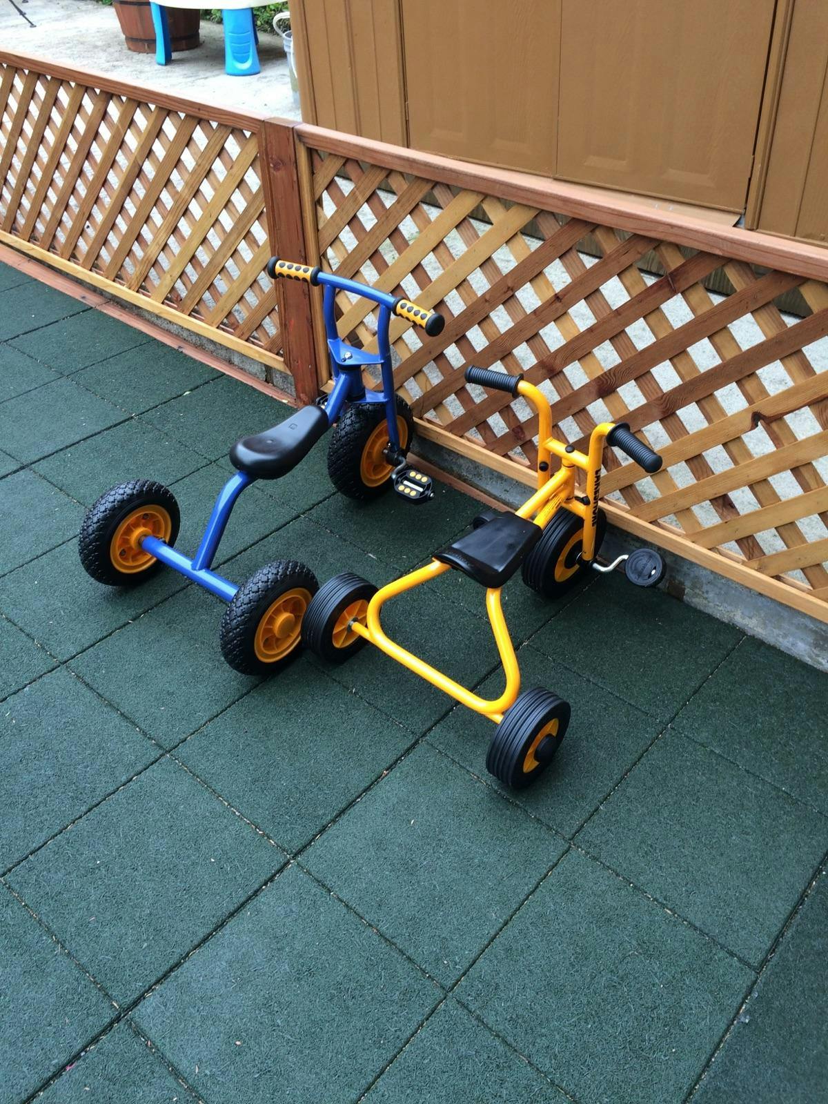 Blue and yellow tricycles on a dark green tiled surface next to a wooden fence