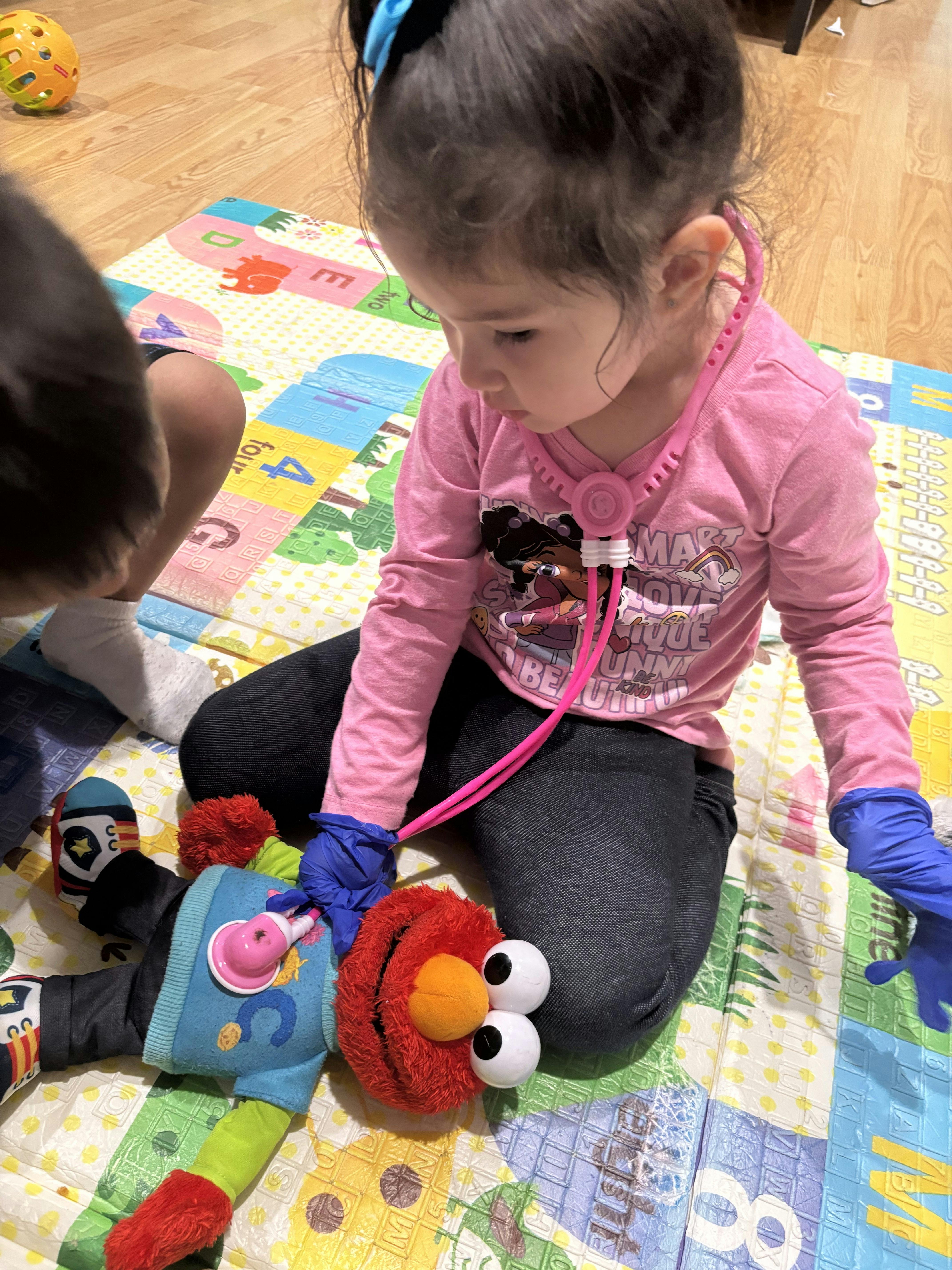 Child wearing blue gloves and a pink play stethoscope, examining an Elmo doll