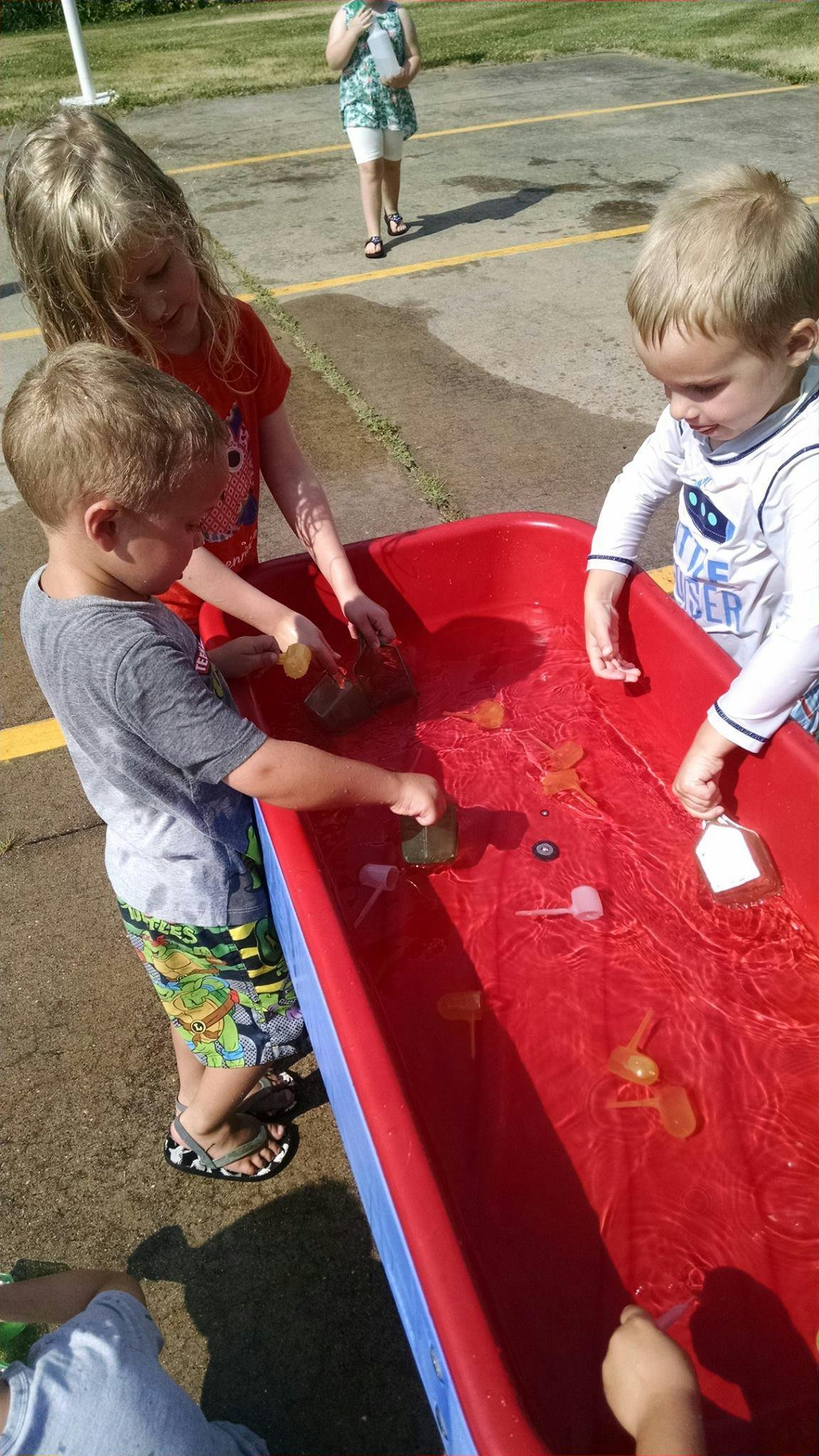 Three children playing with toys in a red water table outdoors