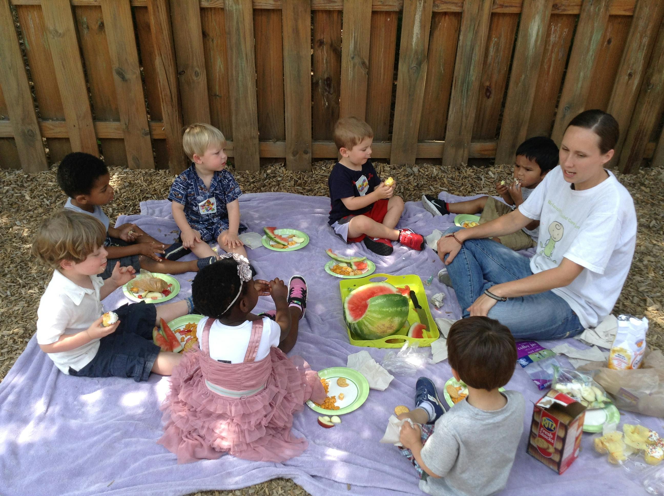 Group of children and an adult eating snacks on a picnic blanket outdoors