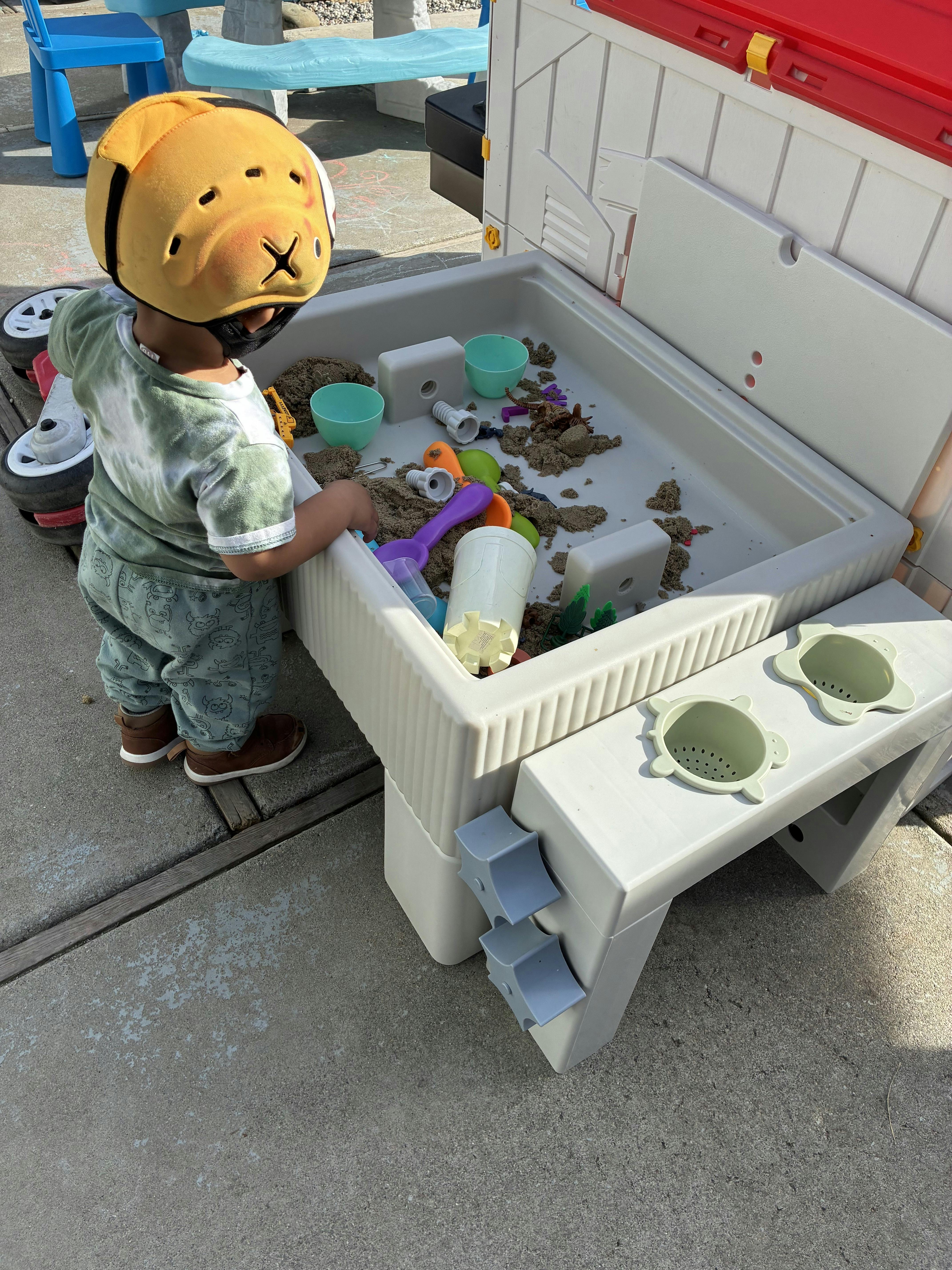 Child in a yellow head protector playing with sand toys in a sandbox