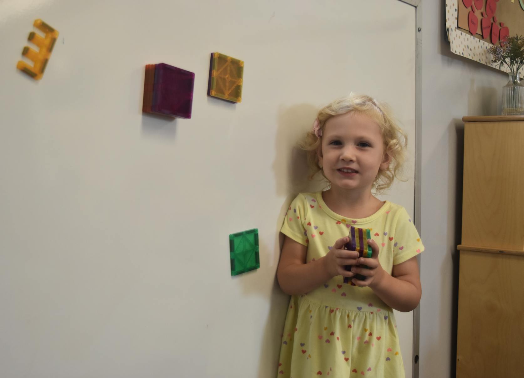 Child holding an object, standing next to a whiteboard with magnetic letters