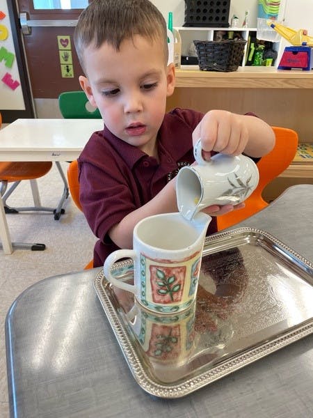 Child pouring from a small pitcher into a mug on a tray