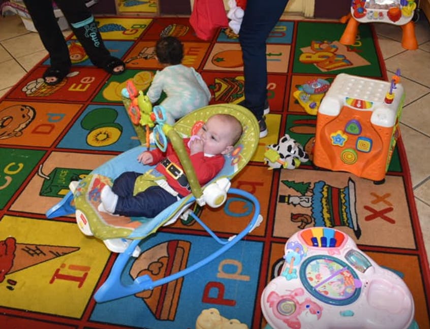 Baby in a bouncer on a colorful alphabet rug with another child and adult legs