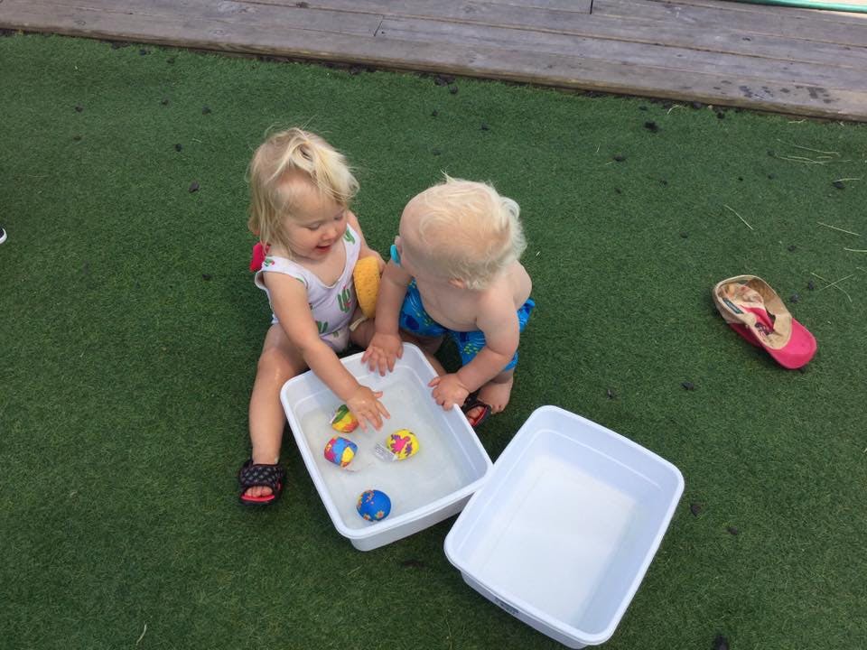 Two children playing with water and toys in a bin on artificial grass