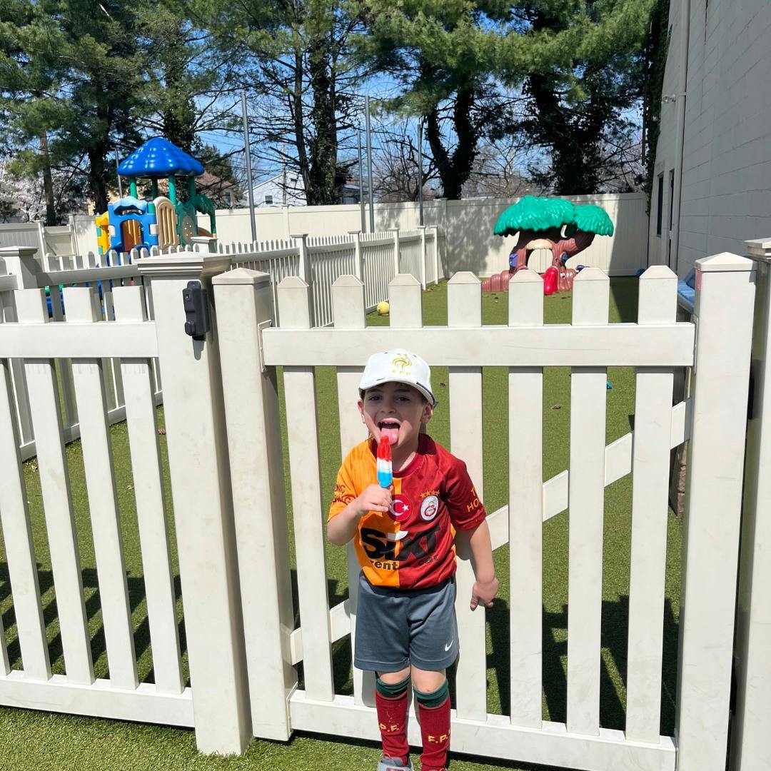 Child in a cap and shirt holding a popsicle, standing by a white fence