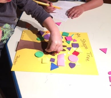 Child placing foam shapes on a yellow paper with "My Shape Tree" written on it