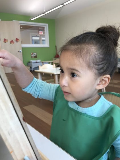 Child in a green smock painting on an easel in a classroom