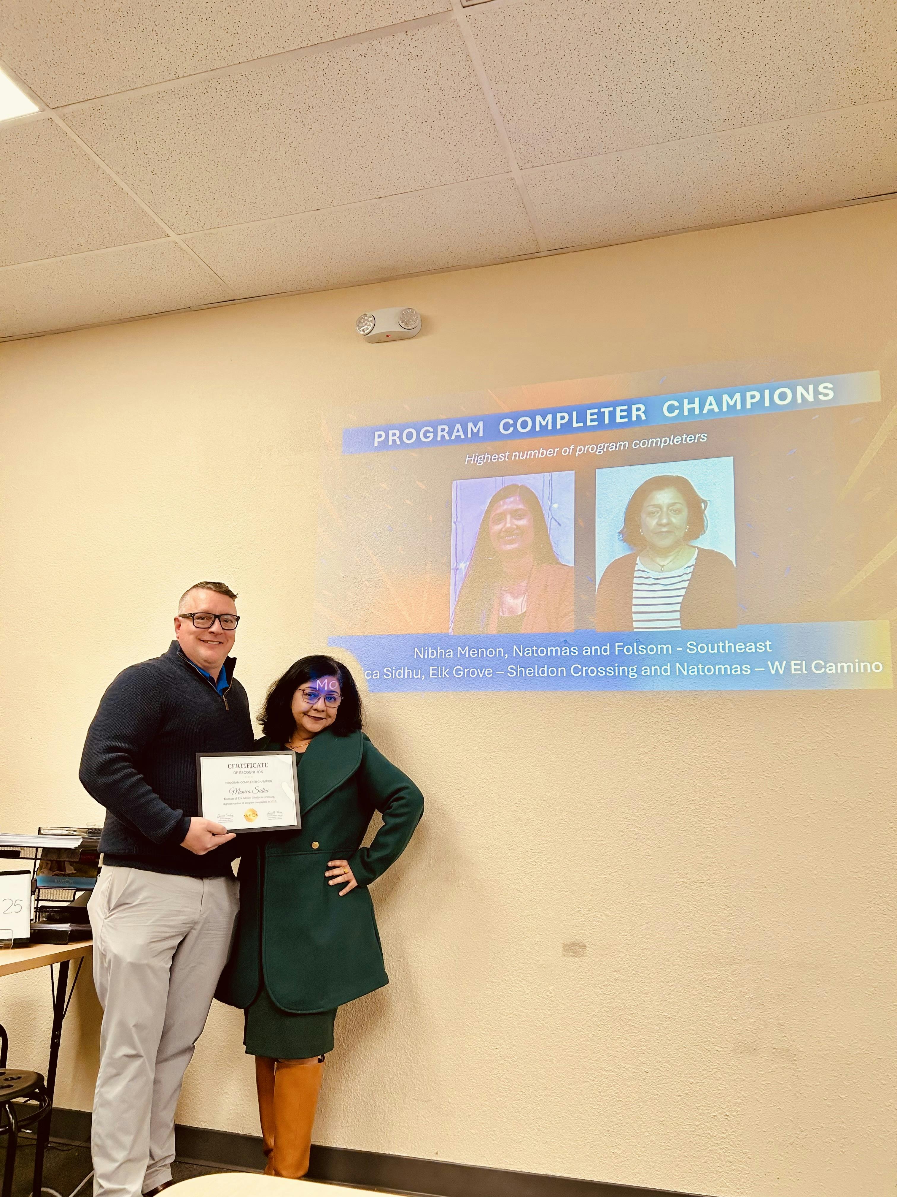Two people standing in front of a projector screen titled Program Completer Champions